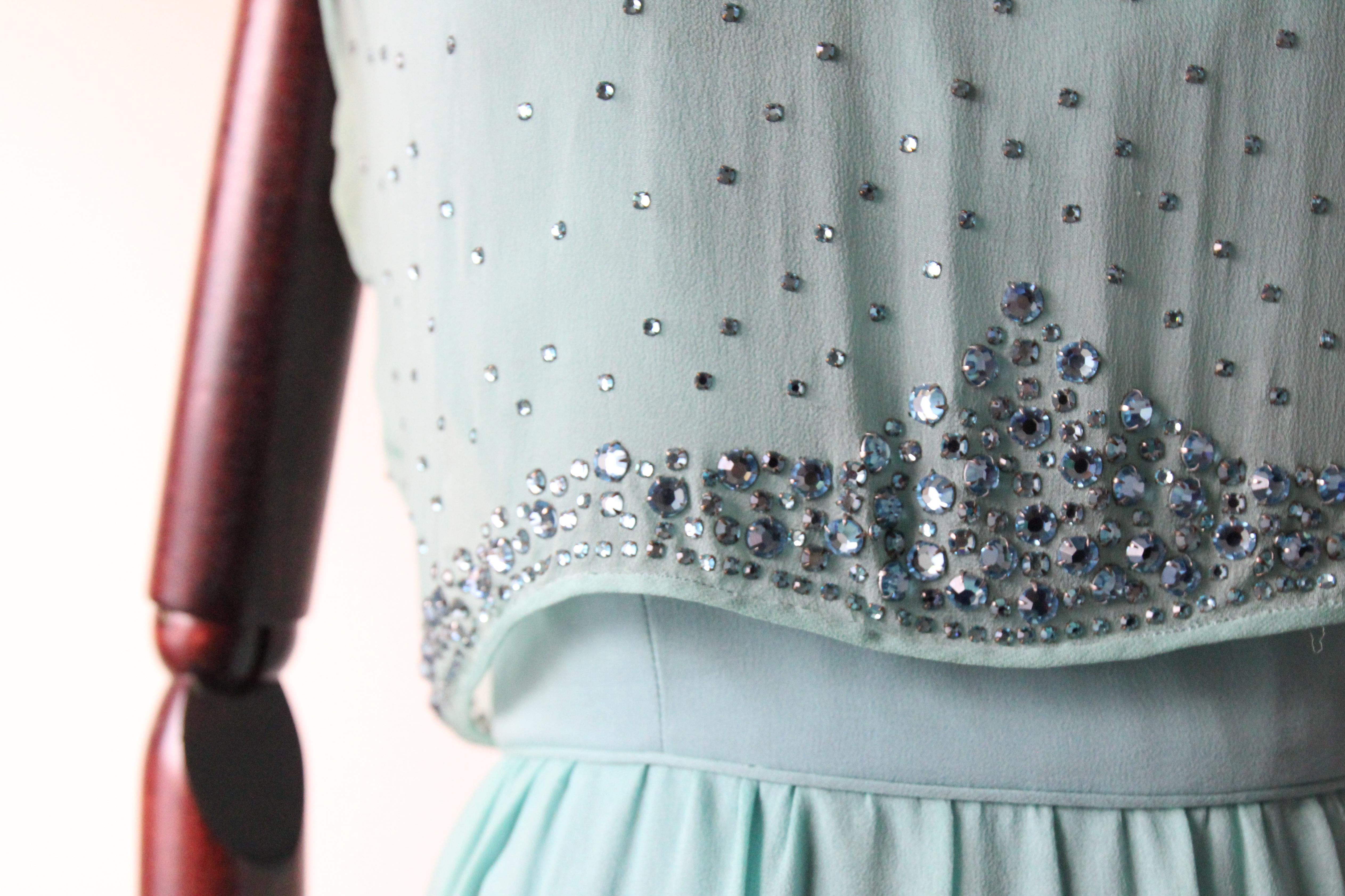Este impresionante vestido de gasa de seda azul Tiffany de los años 60, adornado con pedrería engarzada de color azul hielo y gotas de cristal facetado, es la pieza perfecta para esa ocasión especial. 

El escote redondeado está enmarcado por una