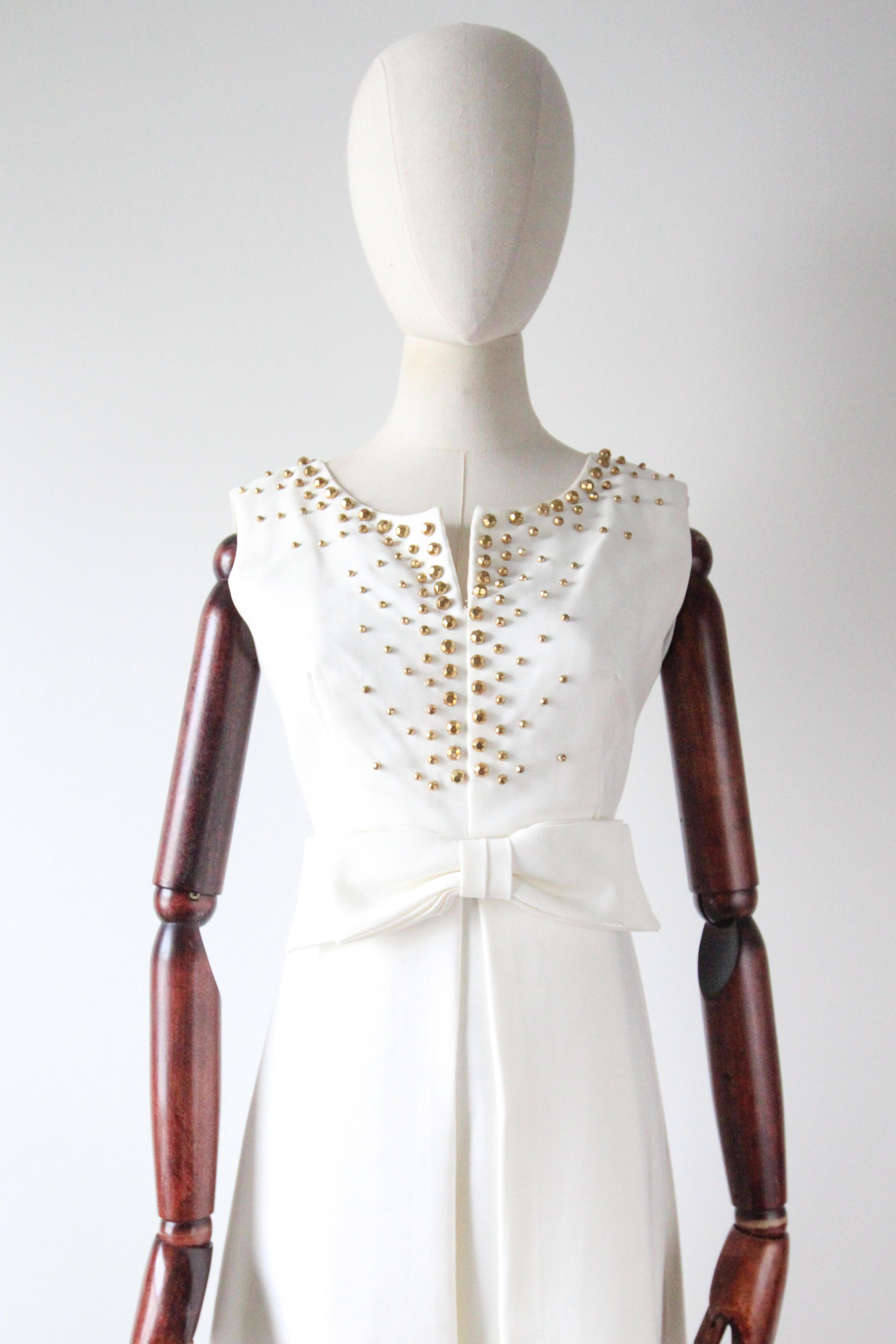 Este icónico vestido blanco de los años 60, acentuado con tachuelas doradas facetadas en forma de garra, en un patrón disperso a lo largo del escote y el corpiño, rematado con un lazo central decorativo, es la pieza perfecta para tu vestuario de
