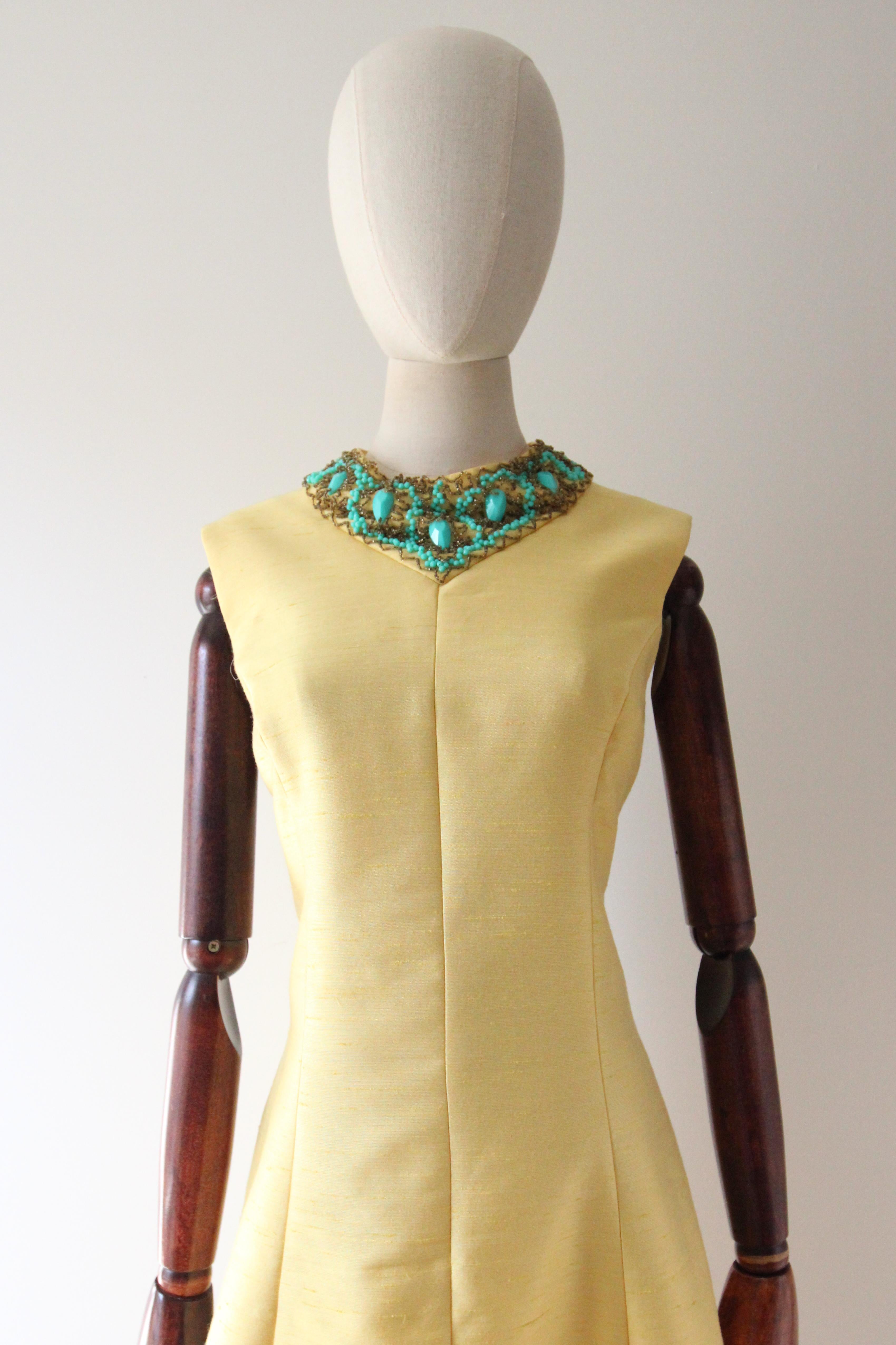 Este vestido original de los años 60, de un vibrante tono amarillo de seda cruda y acentuado con abalorios plateados y azul turquesa, es la pieza perfecta para añadir a tu colección vintage.

El escote redondeado está enmarcado por un detalle