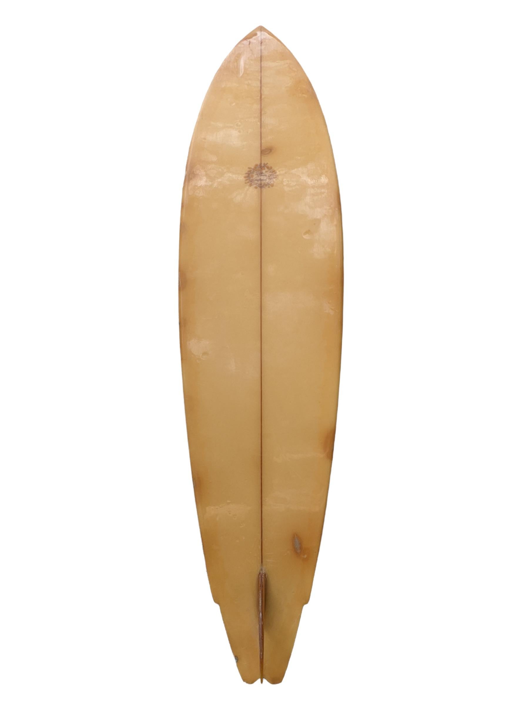 Planche de surf Dick Brewer de la fin des années 70, fabriquée par Bob Ricard. De teinte jaune, avec un design en forme de queue d'hirondelle ailée. Rayures centrales, contour de la planche rayé et aileron simple vitré. Un magnifique shortboard des