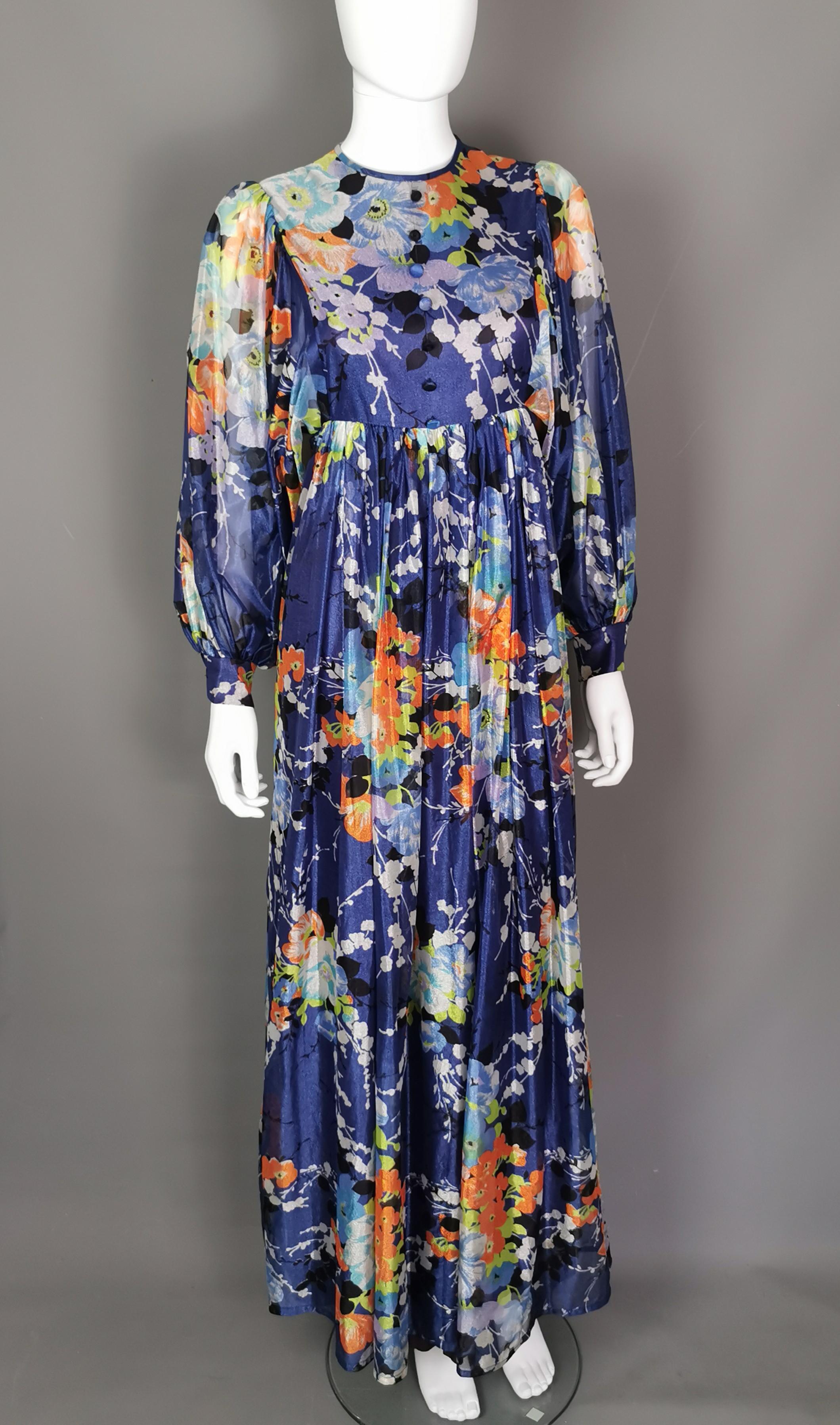 Ein wunderschönes Maxikleid mit Umhang aus den 1970er Jahren in einem leuchtenden Himmelblau.

Dieses Kleid erinnert mit seinem leichten Stoff, dem floralen Muster und dem langen, ausladenden Rock an sonnige 70er-Jahre-Festivaltage.

Es ist aus