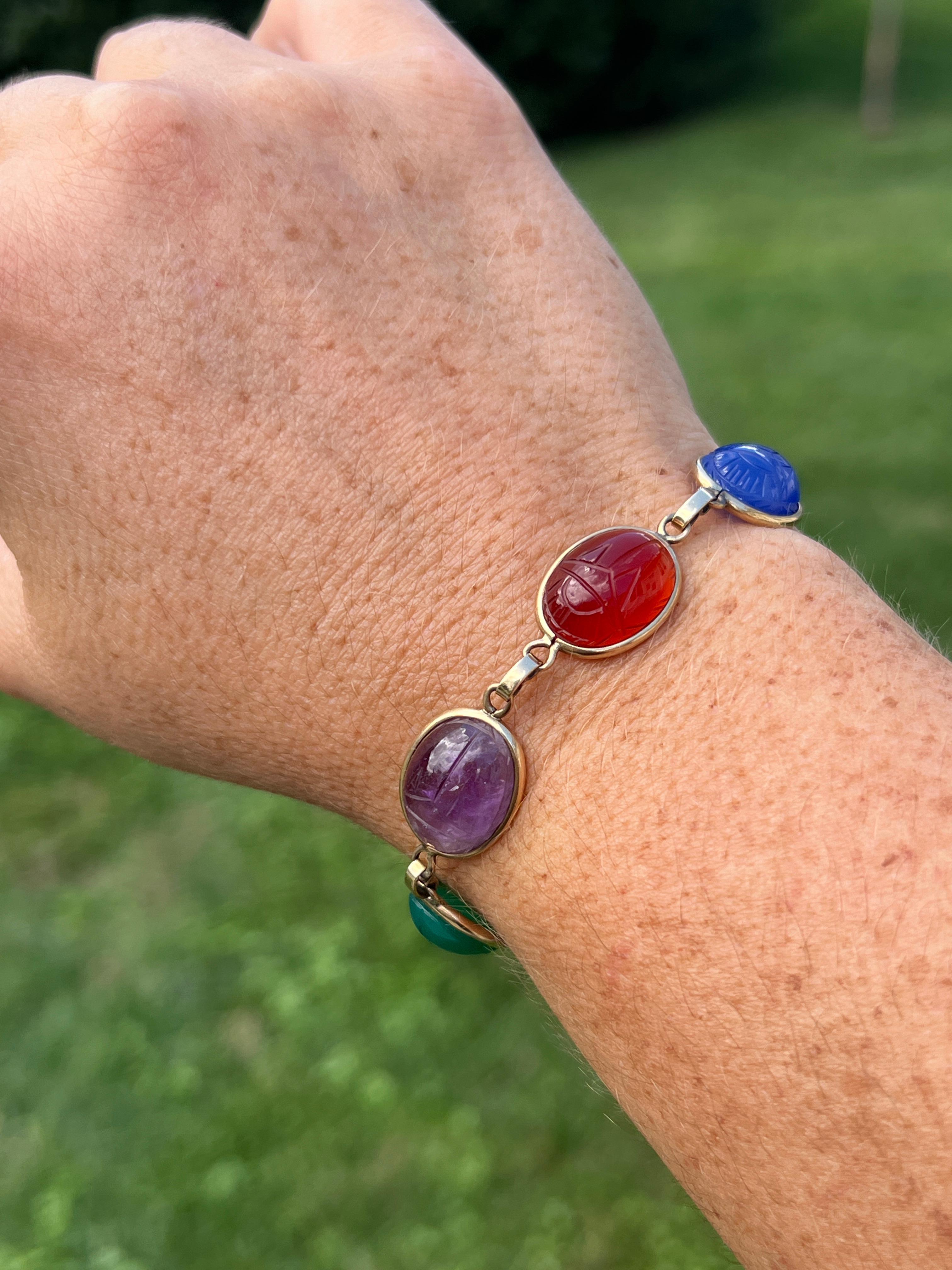 This vintage 1970s bracelet, crafted from 14 karat yellow gold, is a vibrant display of carved scarab gemstones. The bracelet features six oval 13.5mm scarabs, each intricately carved from a different gemstone: citrine, dyed blue chalcedony,
