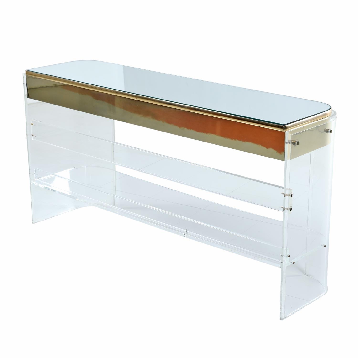 This vintage 1970s neo-deco brass / gold colored Lucite acrylic console exudes glamour. Light bounces off the curved brass trim and refracts through the Lucite. The transparent lower tiered shelf adds practicality and almost disappears, which gives