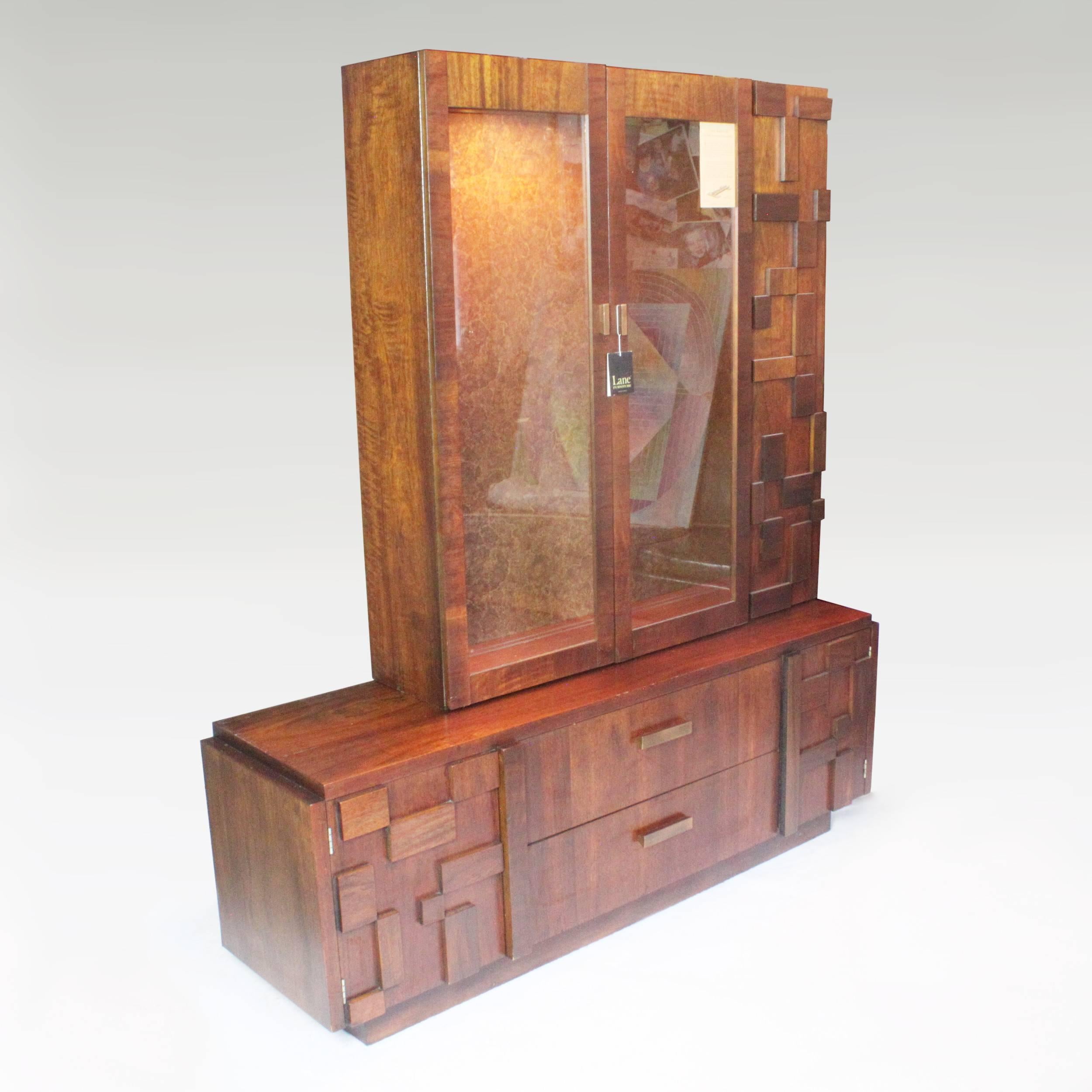This striking piece by lane features applied mosaic blocks motifs that give it an exemplary Brutalist style. Cabinet features walnut veneer, two-door or two-drawer lower cabinet and twin glass door upper cabinet with interior light and two glass