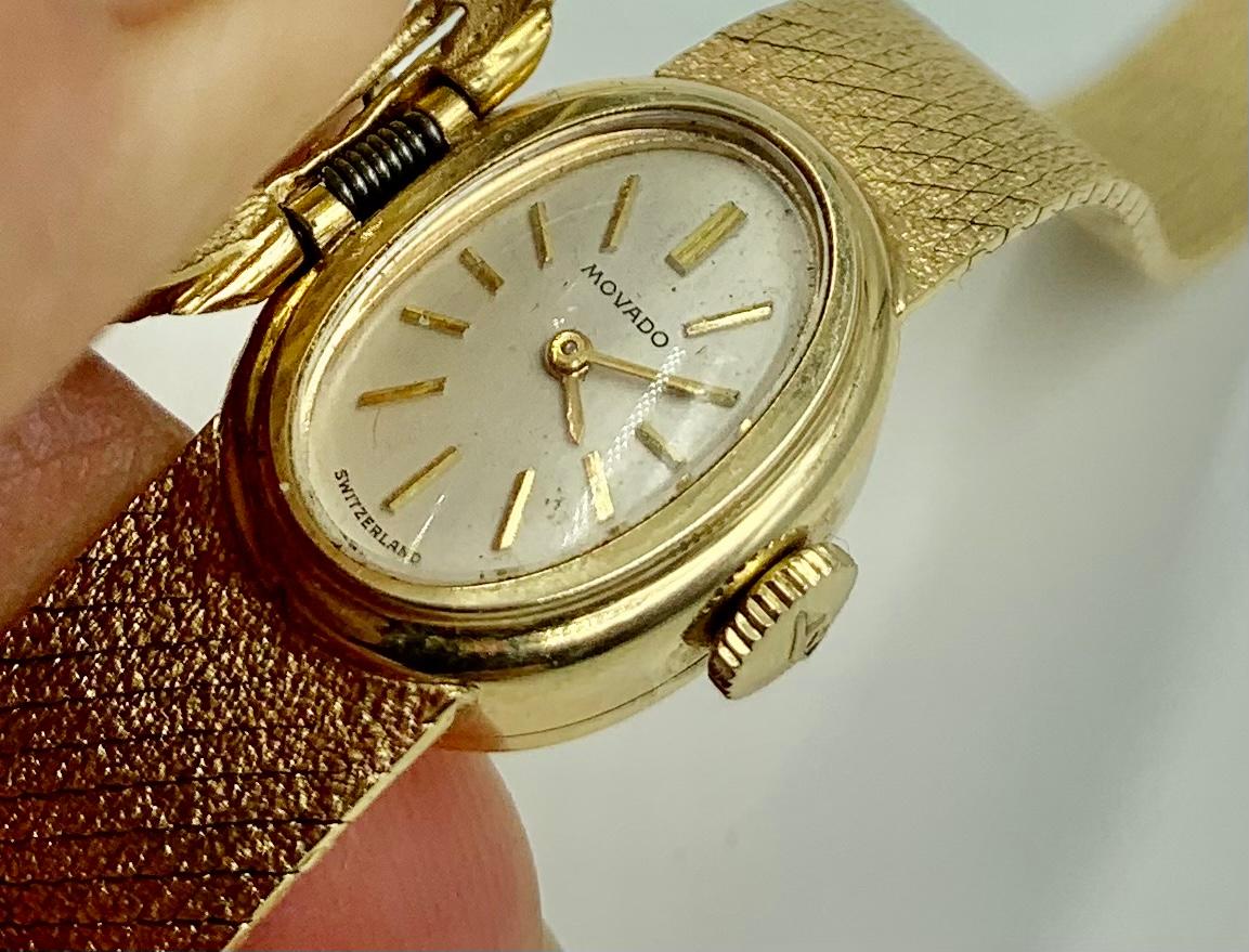 Hidden watch compartment under a spring mechanism rope twist textured oval gold cover with a fine textured gold watch band ending in a double locking clasp. Oval cream colored watch face signed Movado and Switzerland, the back of the watch marked