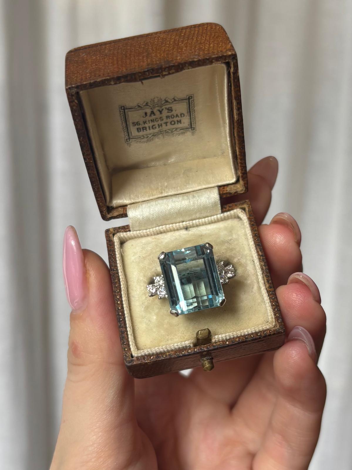 A beautiful vintage 1980's 11.00ct aquamarine and diamond cocktail ring set in platinum.

The piece is centrally set with a sparkly emerald cut aquamarine; which is securely four law set to centre. The aquamarine is sided by three round brilliant
