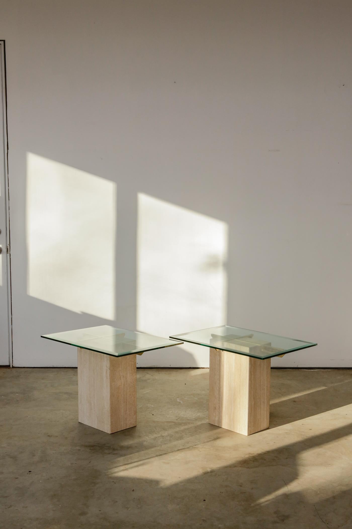 Vintage 1980s Artedi Italian Travertine and brass side tables. Sold Individually. A pair of sleek, chic side tables with two brass supports fitted inside the travertine marble base. Two glass tops, two travertine stone bases, two tables you need in