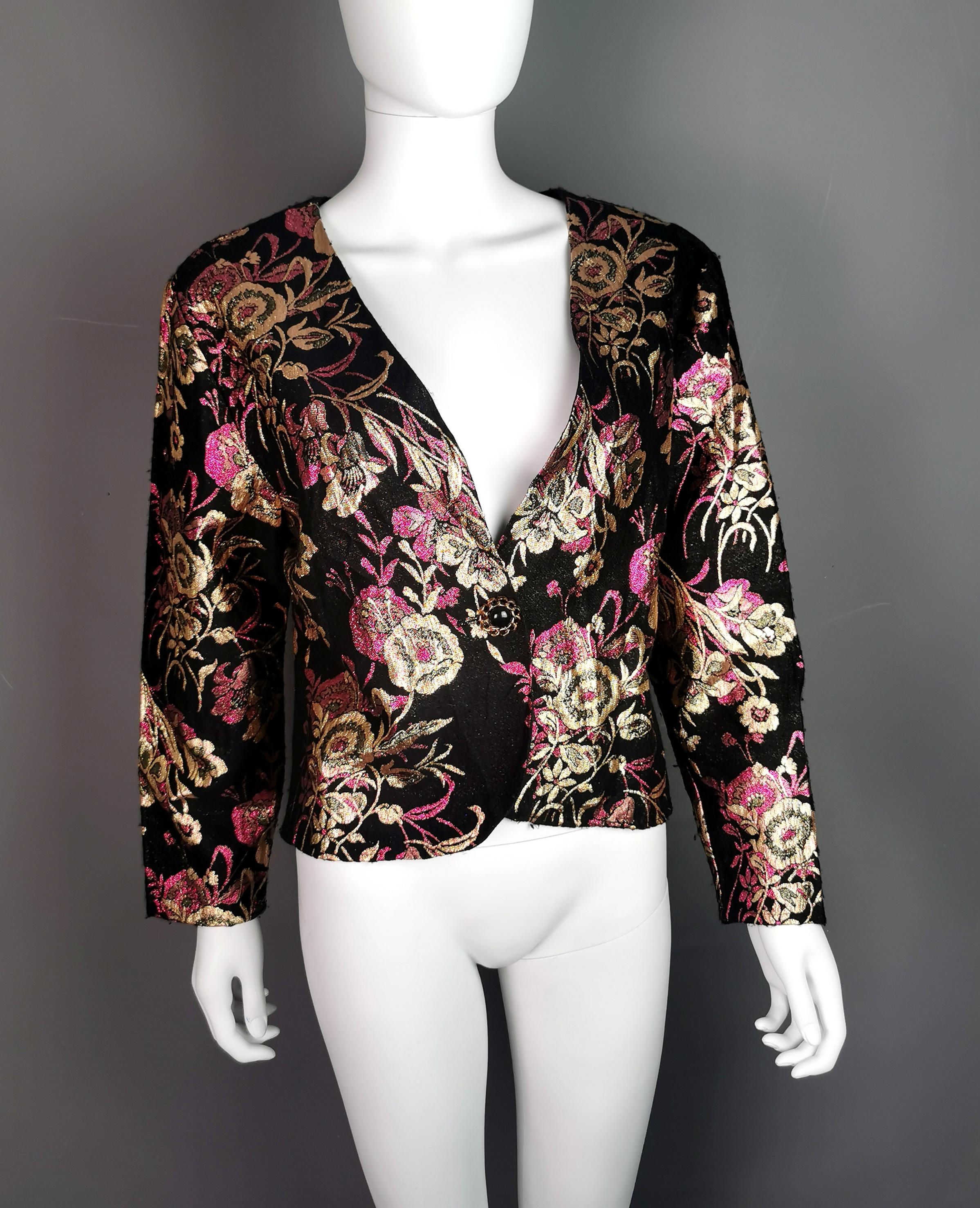 A unique vintage c1980s cropped length Brocade jacket.

It has a black ground with metallic gold and fuschia pink floral Brocade.

The jacket is a shorter length with long sleeves and fastens at the fringe with a single plastic button.

A perfect