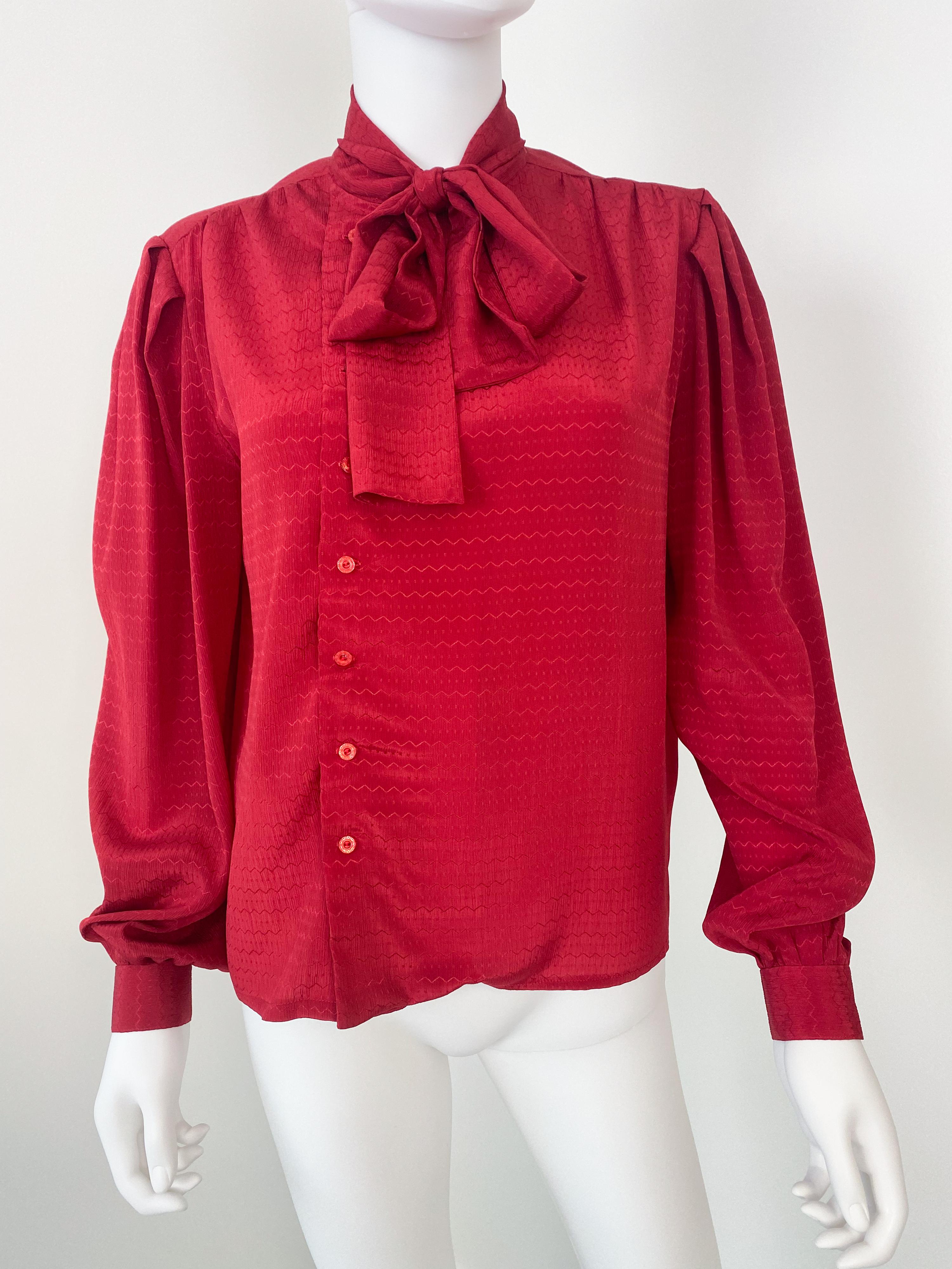 Splendida camicetta vintage anni '80 in poliestere setoso con fiocco a foulard. Tessuto di colore rosso vivo con motivo a righe geometriche tono su tono. Le spalle e i polsini con bottoni presentano un'ampia plissettatura per dare volume.

Marchio: