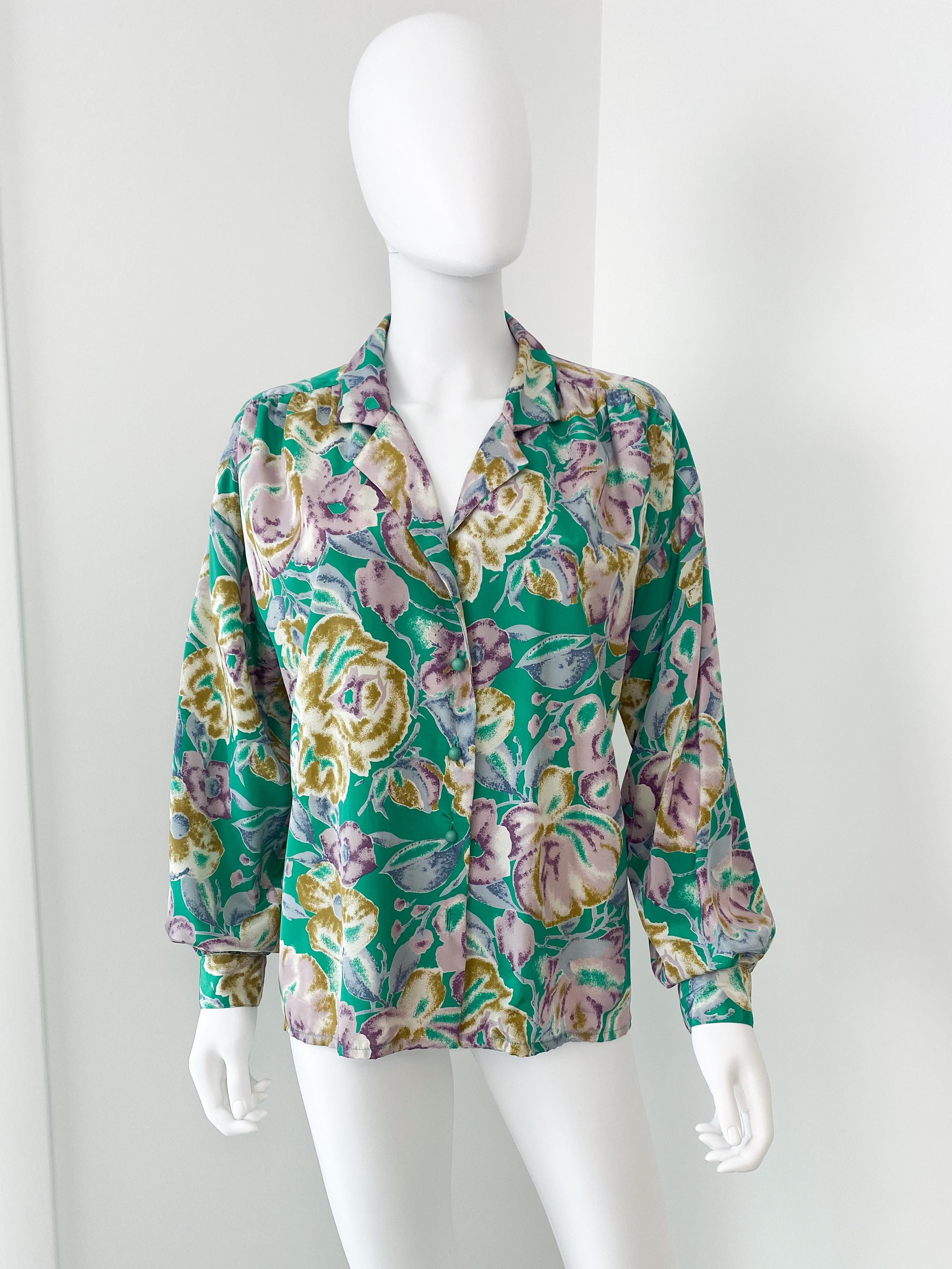 Deliziosa camicetta vintage anni '80 in poliestere setoso con sciarpa a fiocco aggiuntiva per creare look versatili. Tessuto di colore dominante verde con un grande motivo floreale a contrasto color lavanda, oro e bianco. La plissettatura sul retro