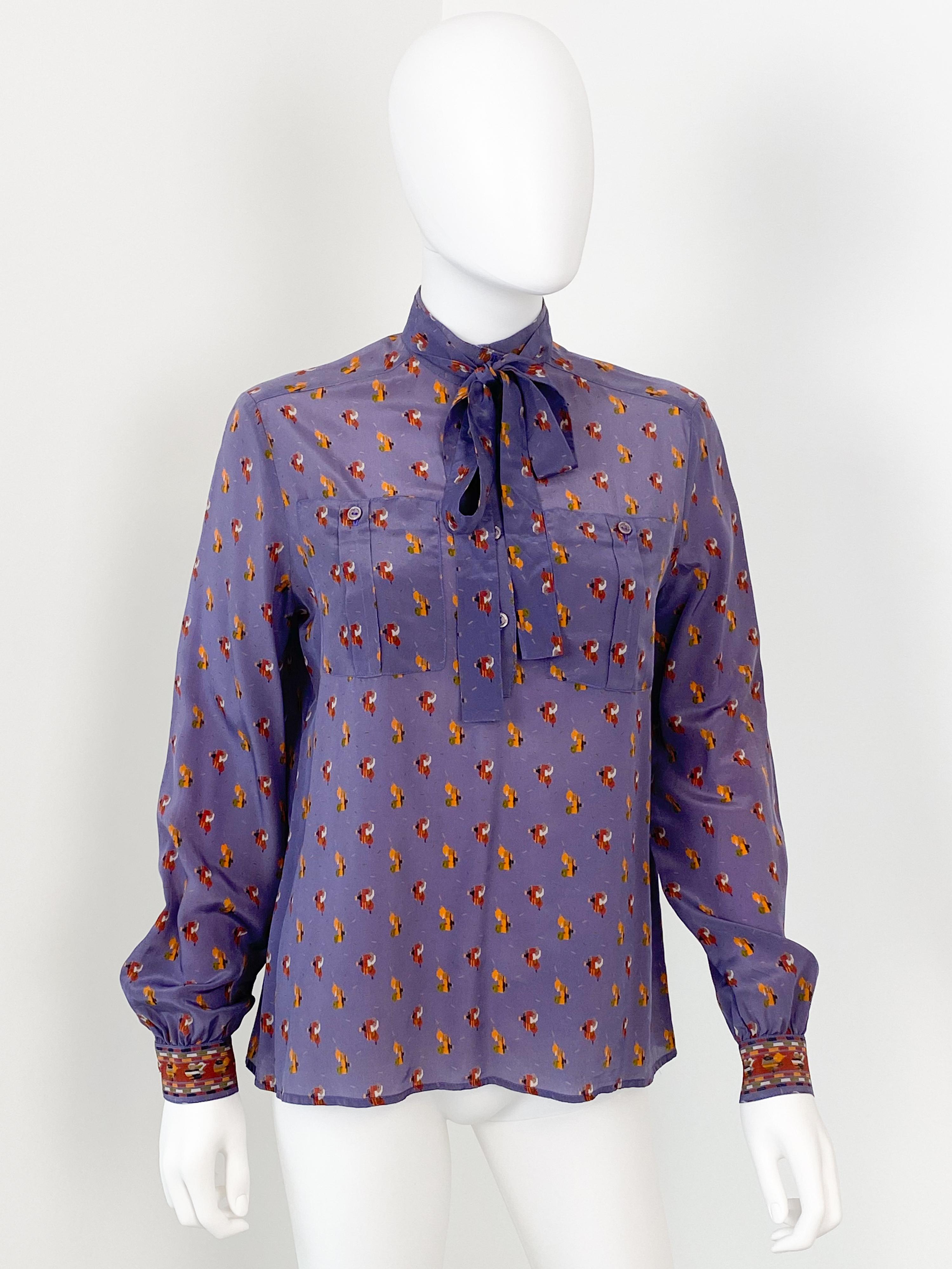 Wonderful vintage 1980s silky polyester blouse in a peasant-style silhouette. Bright purple color fabric with print pattern in orange and burgundy colors. The pullover construction has a partial button front with a tab collar and two pockets. With