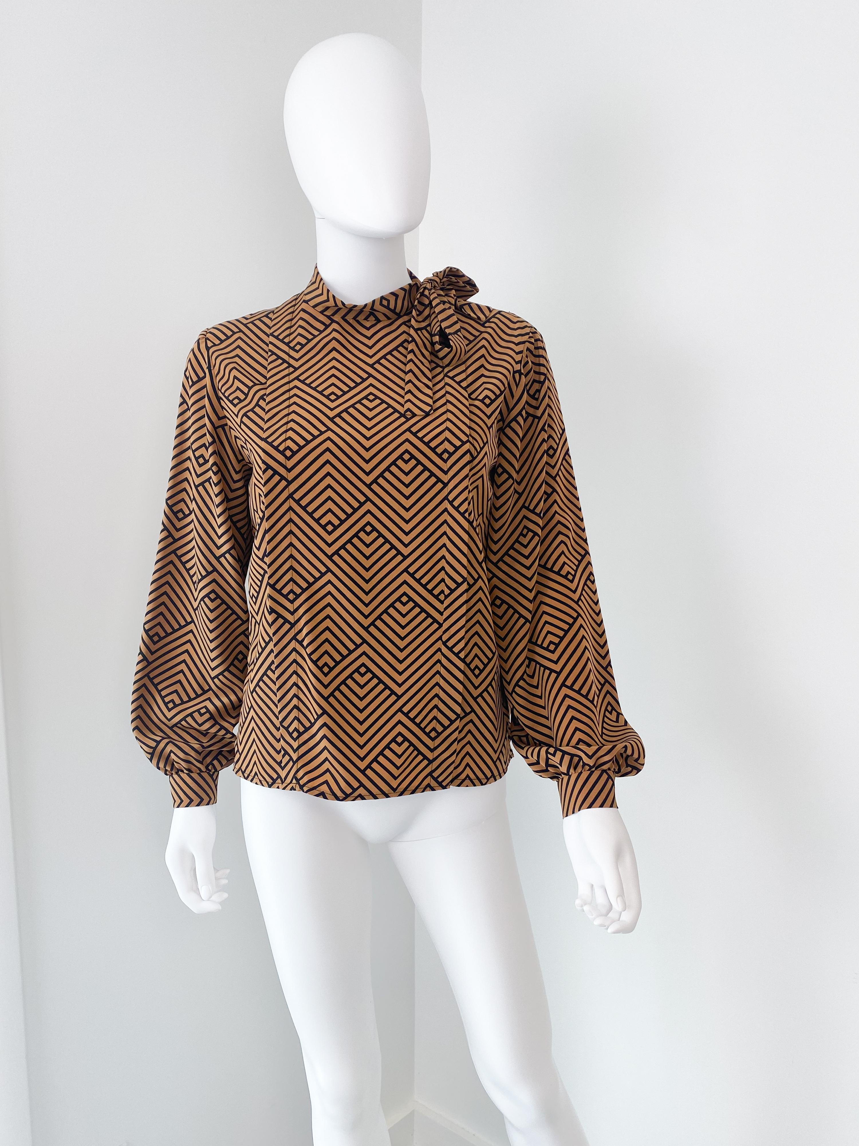 Splendida camicetta vintage anni '80 in poliestere setoso con fiocco a foulard. Tessuto vibrante color sidro arancione con motivo geometrico a zig-zag nero. I polsini con bottoni presentano un'ampia plissettatura per dare volume. Il fiocco deve