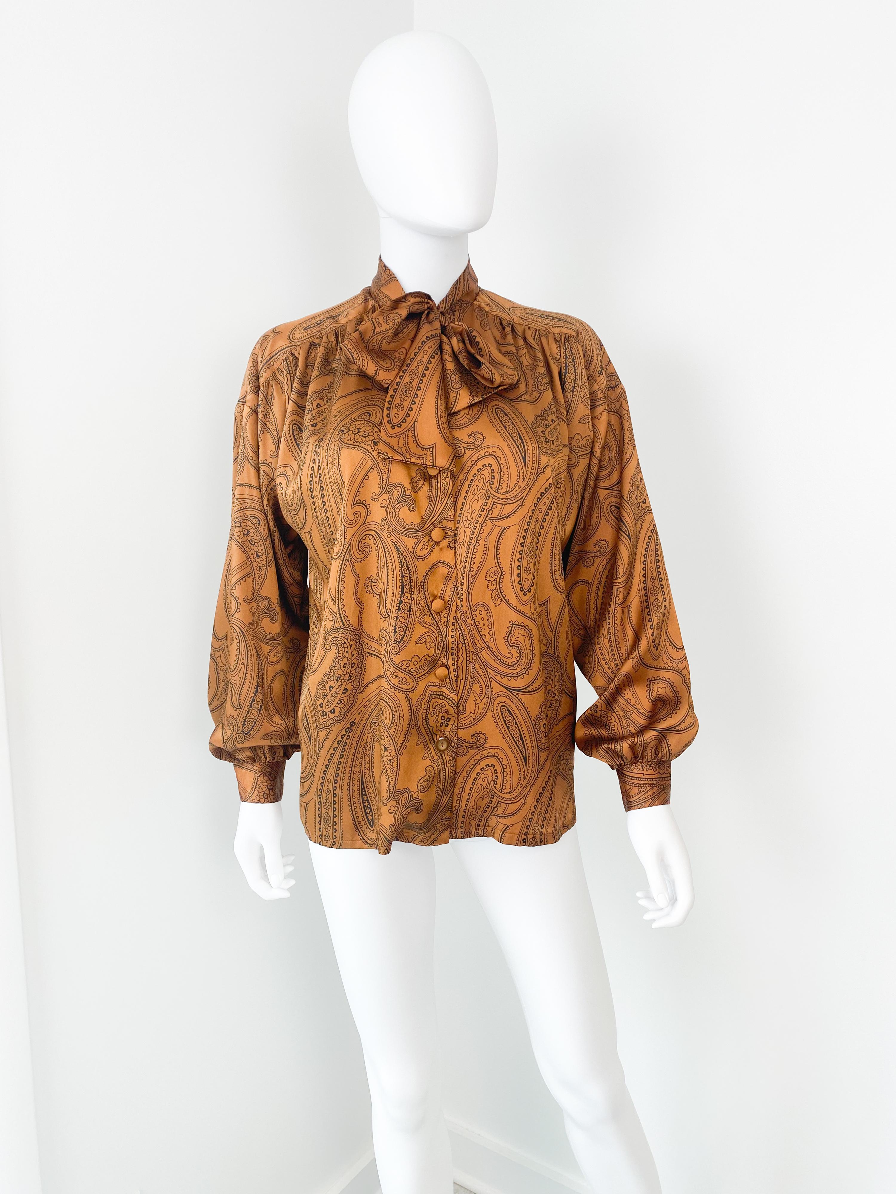 Splendida camicetta vintage anni '80 in poliestere setoso con fiocco a foulard. Tessuto vibrante color arancio zafferano con motivo paisley nero. La plissettatura sul davanti, sul retro e sui polsini con bottoni garantisce volume. L'ampiezza della