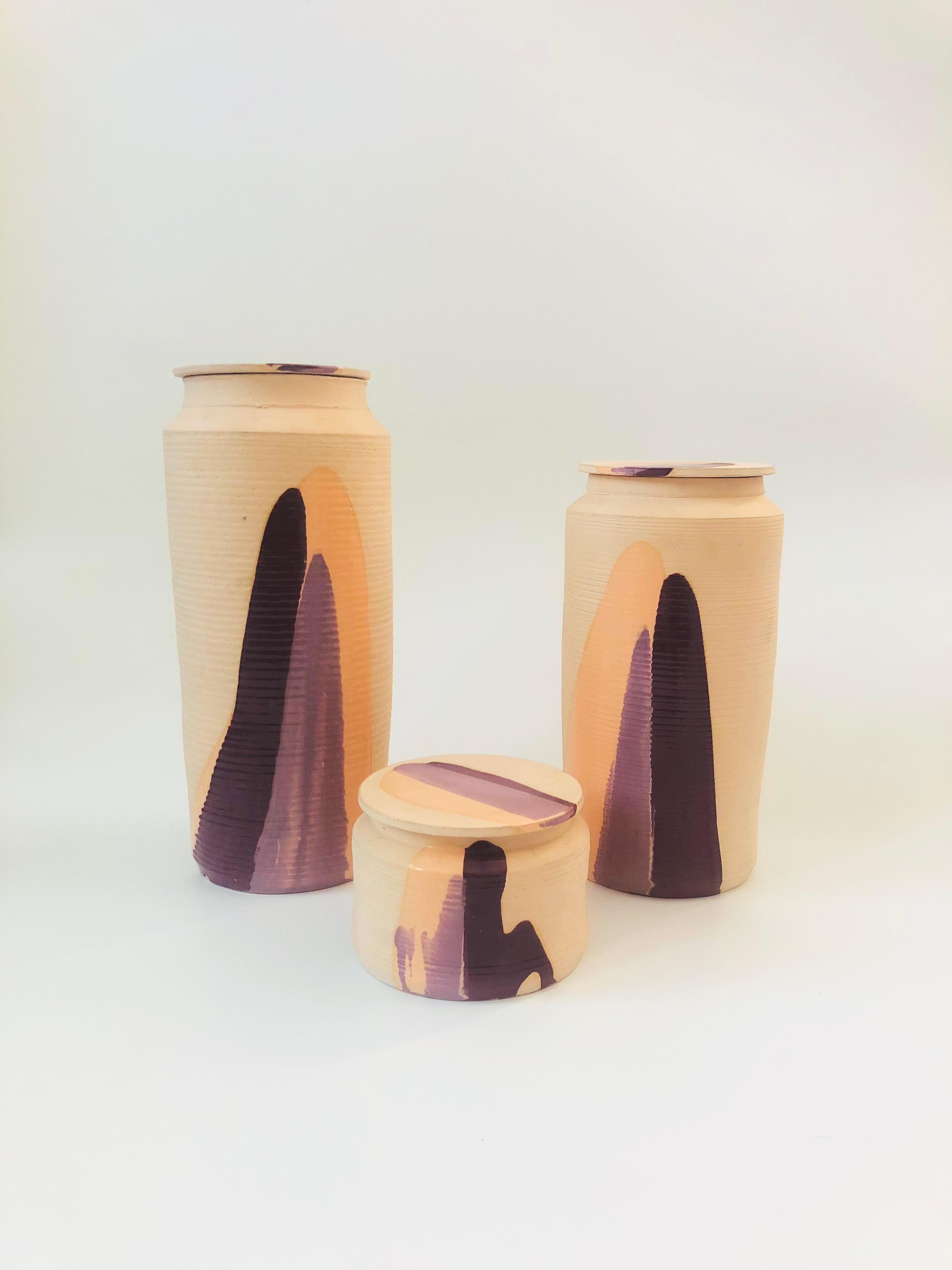 A set of 3 vintage 1980s studio pottery canisters in graduated sizes. Nice simple cylinder shapes. Made of unfinished clay in a pale beige color that has been decorated with high gloss glazes in purple hues. Each is signed 