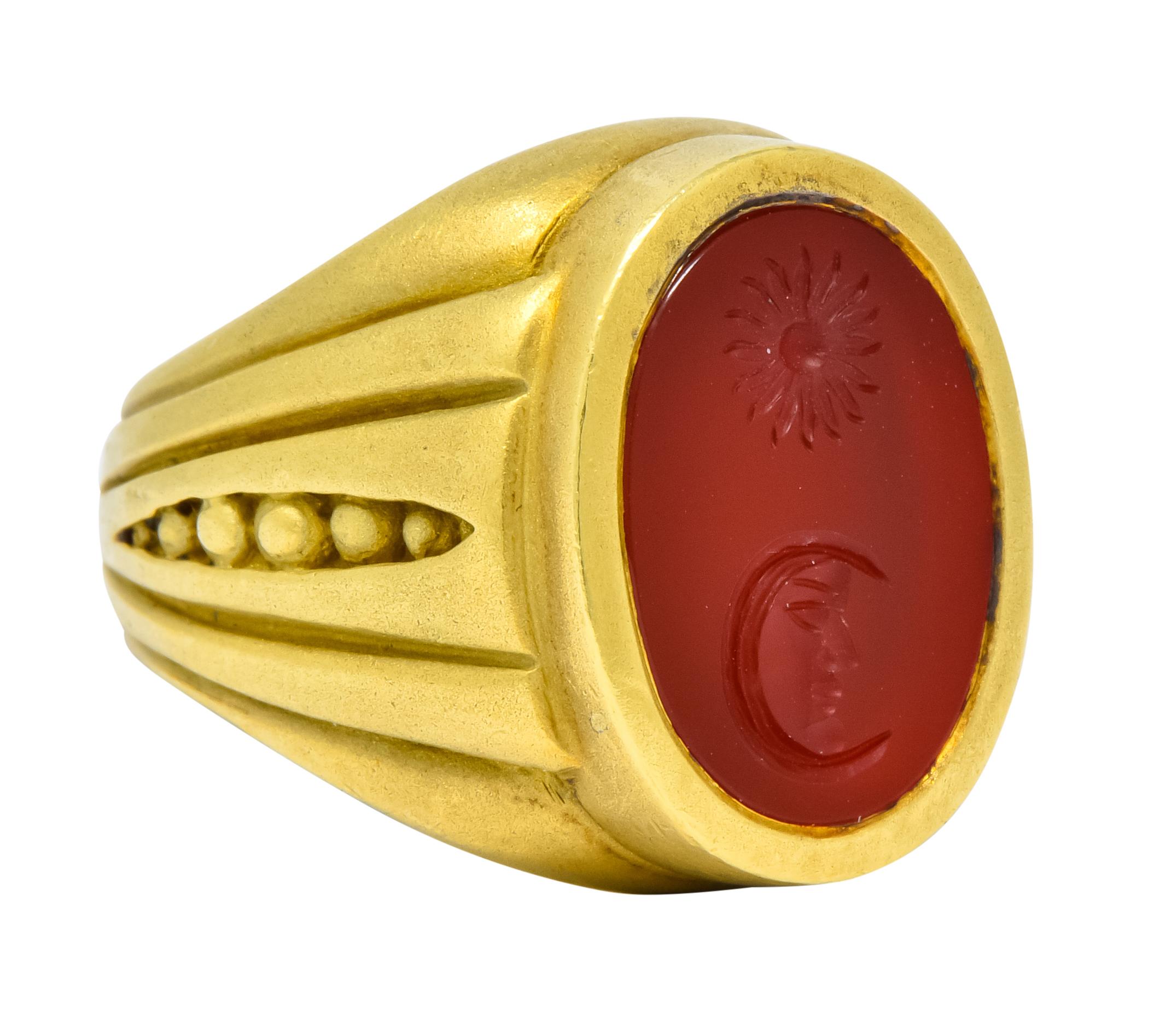 Centering an oval cut tablet of carnelian measuring approximately 5/8 x 7/16 inches, translucent and a vibrant red

Deeply carved with a sun and man-in-moon motif

Bezel set in a matte gold signet style mounting with ribbed shoulders 
centering a