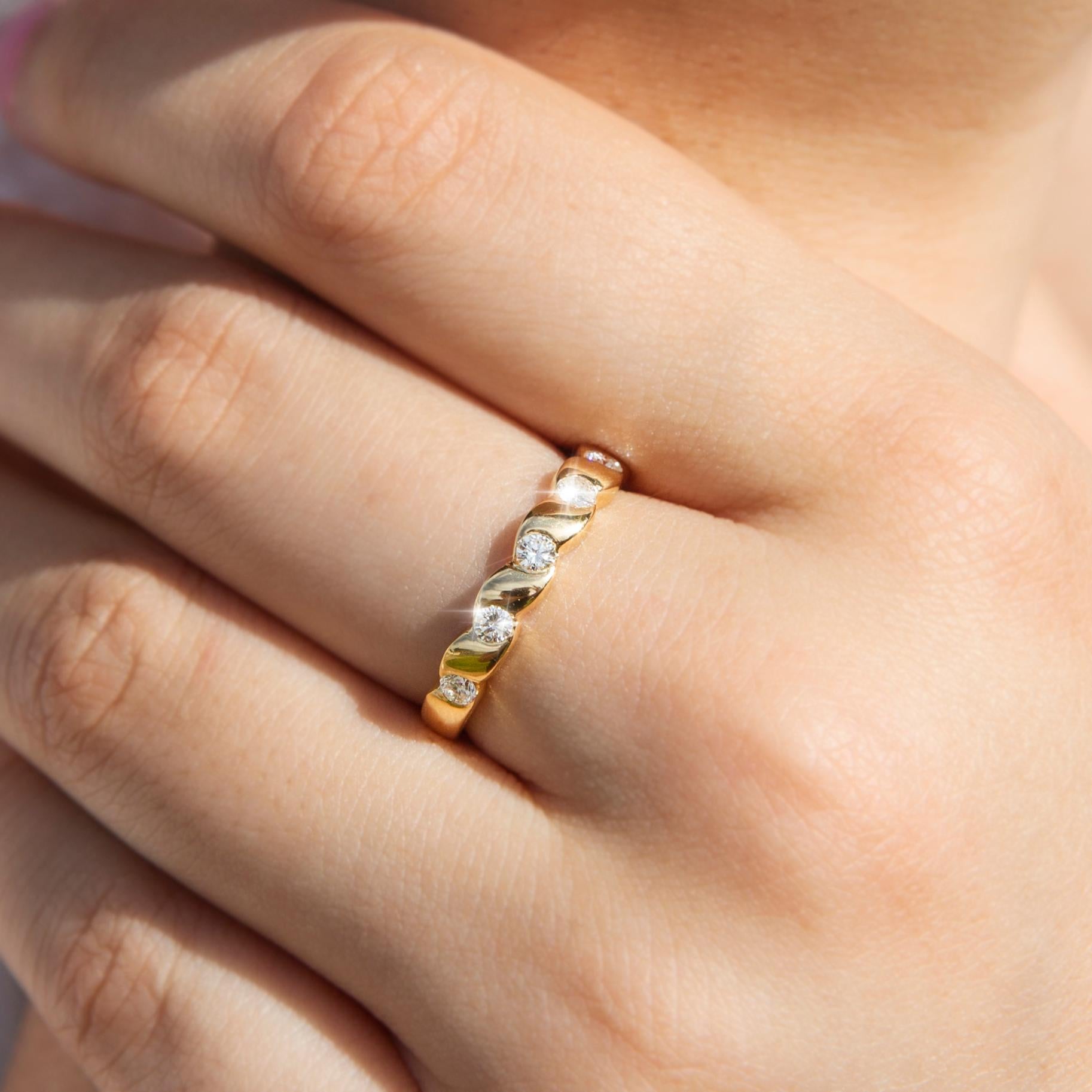 Forged in 18 carat yellow gold, this lovely diamond band ring is a delightful balance of simplicity and elegance. The front of the yellow gold band holds a shimmering row of seven round brilliant cut diamonds set between wave-like bar settings. We