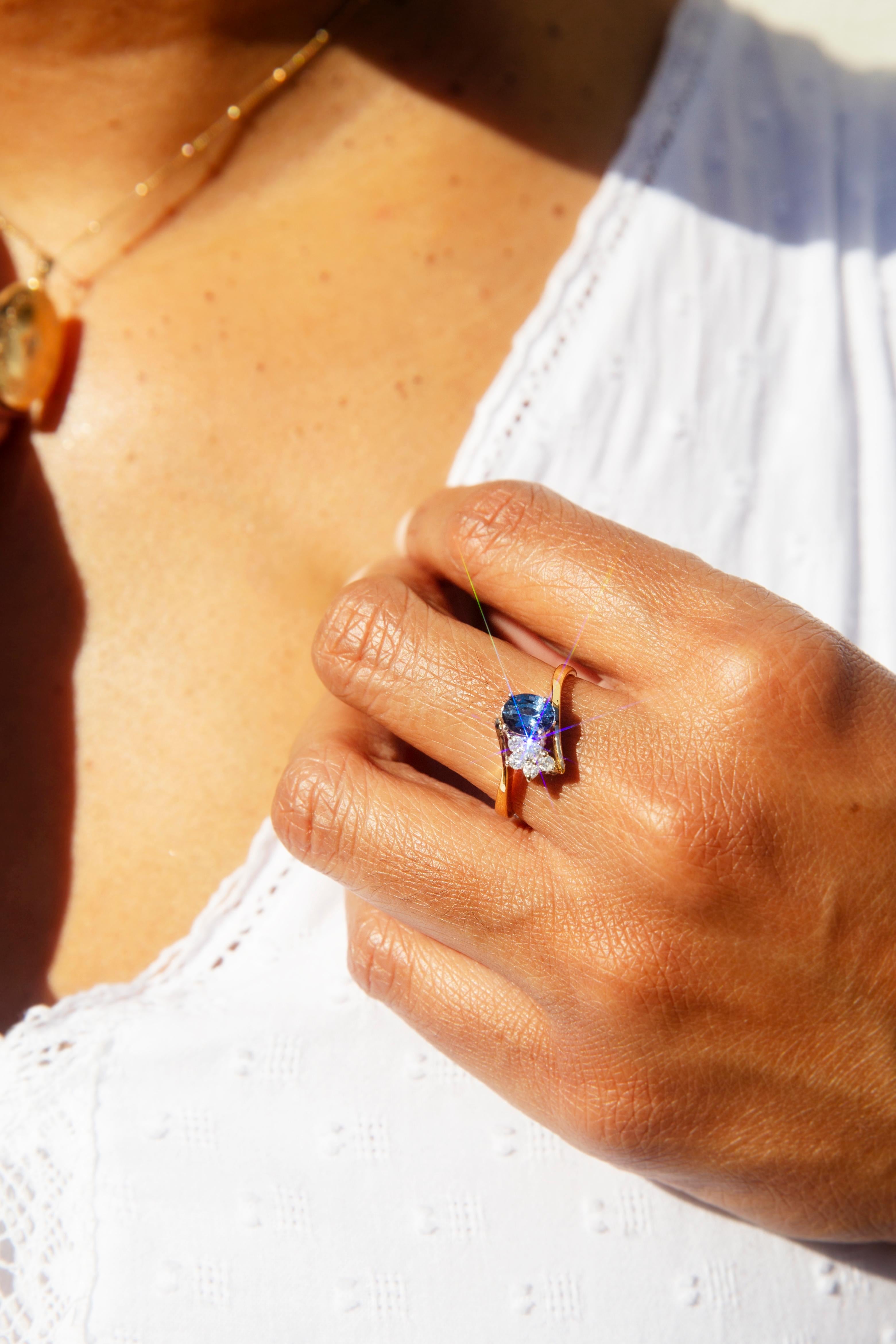 Crafted in 18 carat yellow gold, this charming vintage ring, circa 1990s, features an oval deep blue Ceylon-type sapphire nestled with an adorable cluster of five round brilliant cut diamonds between graceful crossover shoulders. Her name is The