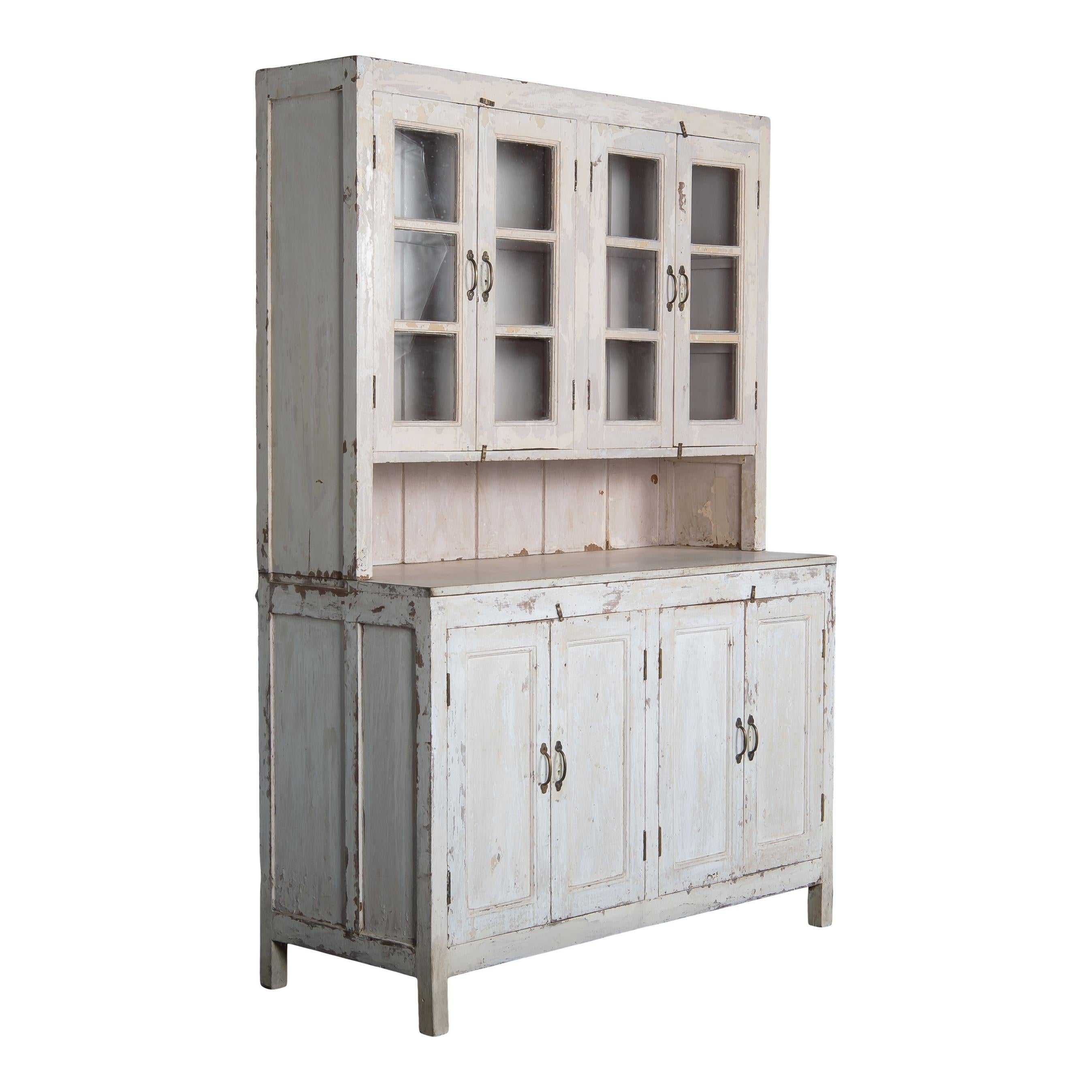 Une armoire classique en bois vintage avec des armoires vitrées, des armoires couvertes et un espace pour le comptoir. Le bois a été peint en blanc qui s'est décoloré et écaillé avec le temps, ce qui lui donne un beau charme vintage. Le haut et le