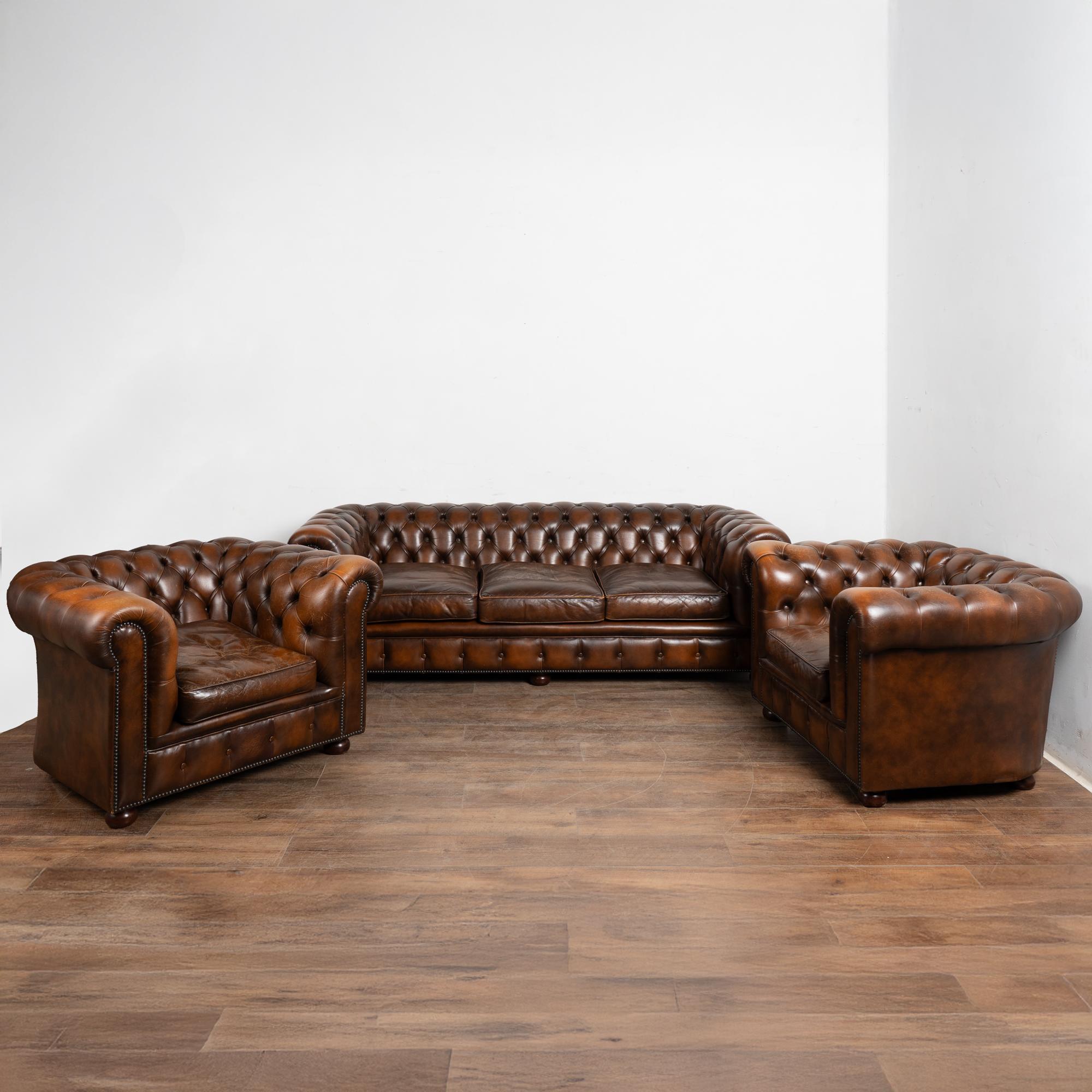 Conjunto de asientos de estilo Chesterfield vintage que incluye un sofá y dos sillones club a juego. Tapicería de piel con mechón marrón, remates de clavos y patas de madera. El desgaste visible incluye rozaduras, arrugas y abrasiones superficiales
