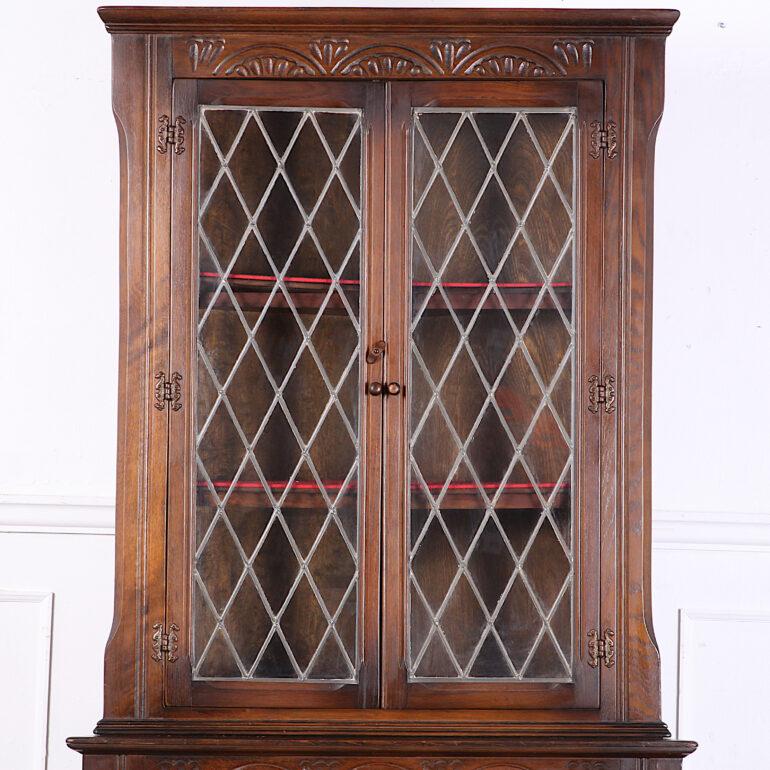 An English mid-20th century standing corner cabinet in oak, the upper section for display behind diamond pattern leaded glass, the lower cabinet with paneled oak doors. Carved details to crown and frame. Nice original finish with warm patina. 

 