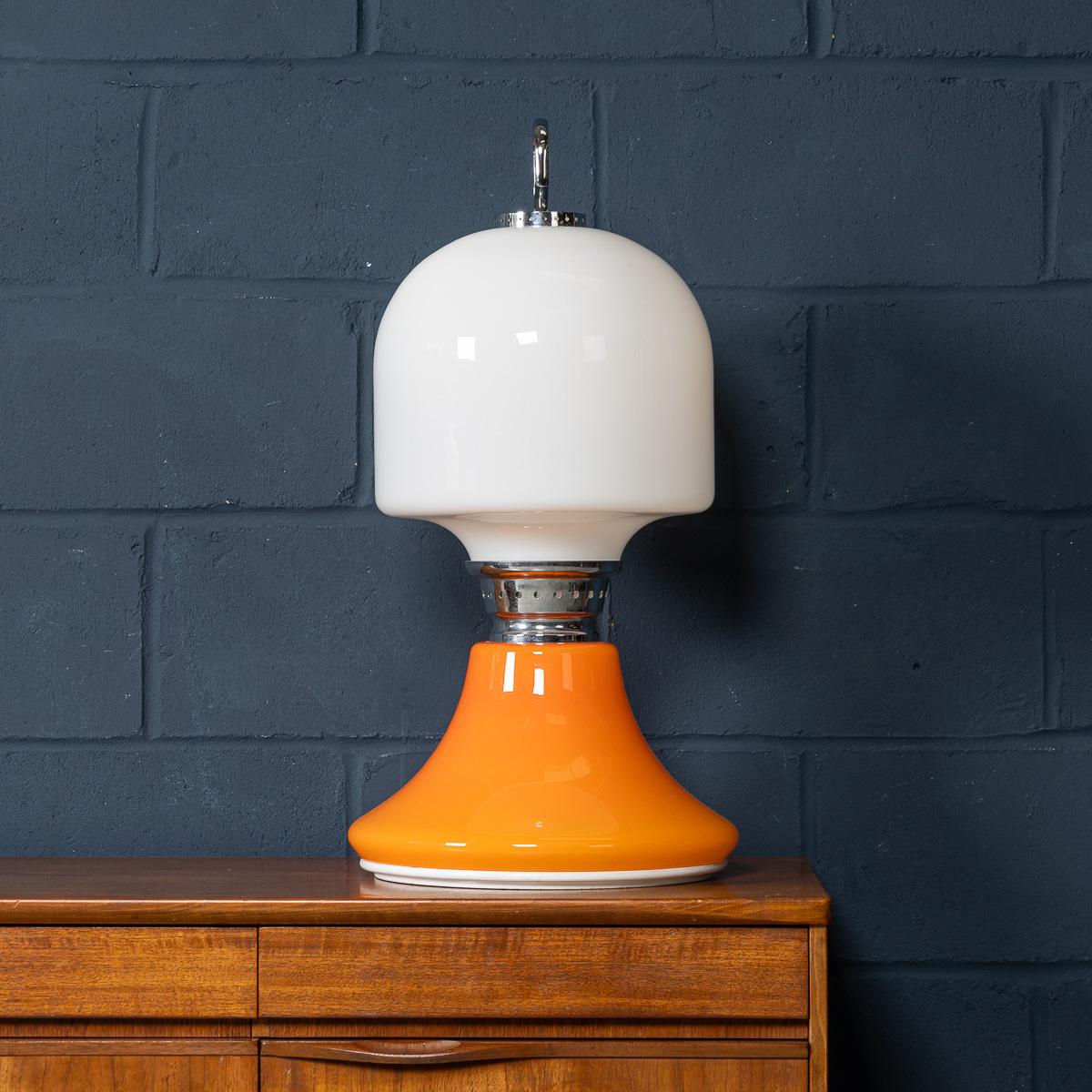 An impressive 20th Century glass table lamp made in Murano, Venice, Italy, around 1970. Crafted in white and rich orange hand-blown glass, the lamp illuminates from both the top and the base, casting a warm and ambient glow. Chrome detailing adds a