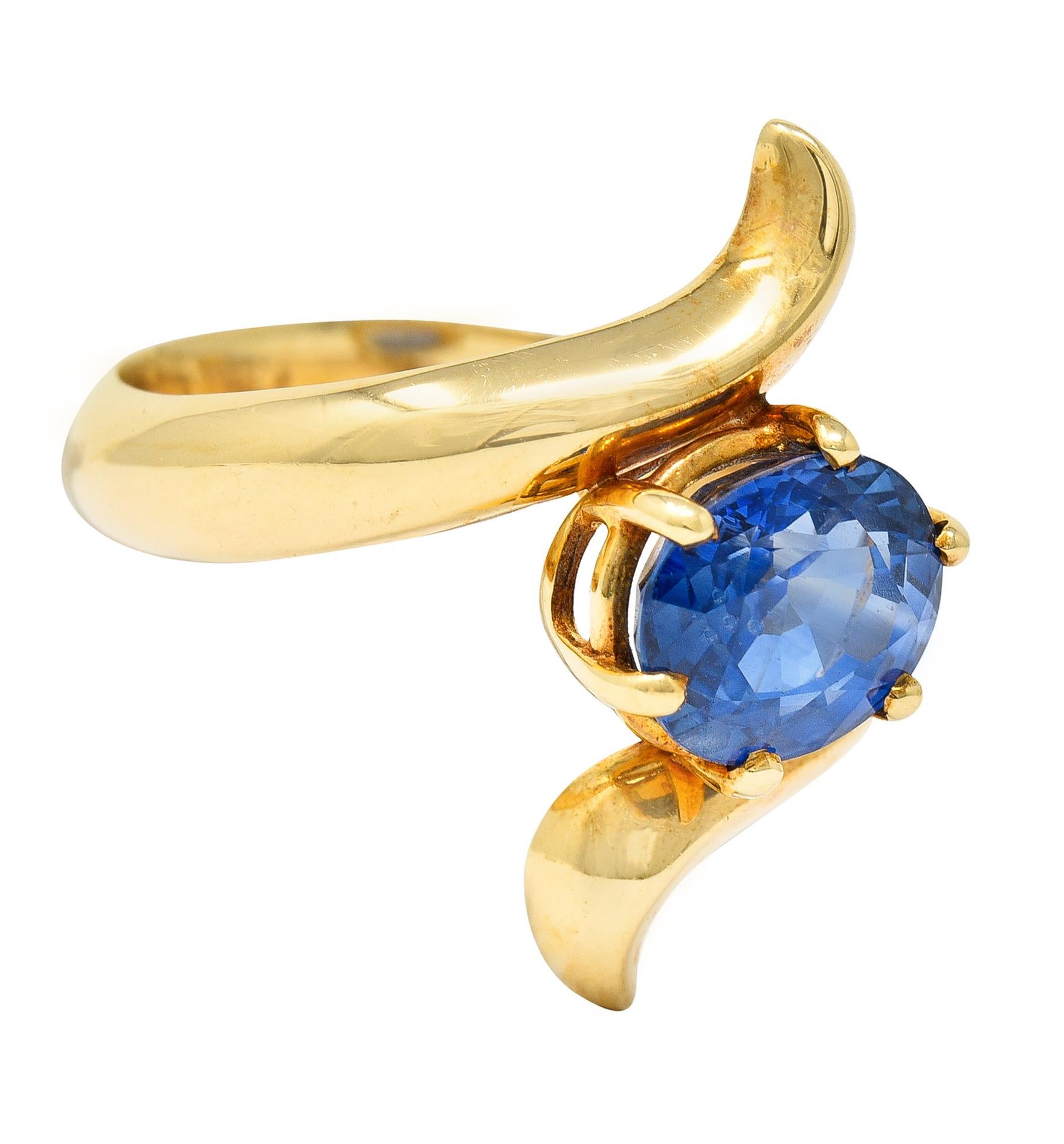 Bypass style ring centers an oval cut sapphire weighing approximately 2.25 carats. Basket set East to West, eye clean, with violetish blue color. Flanked North and South by stylized and scrolling gold tendril shoulders. With maker's mark and stamped