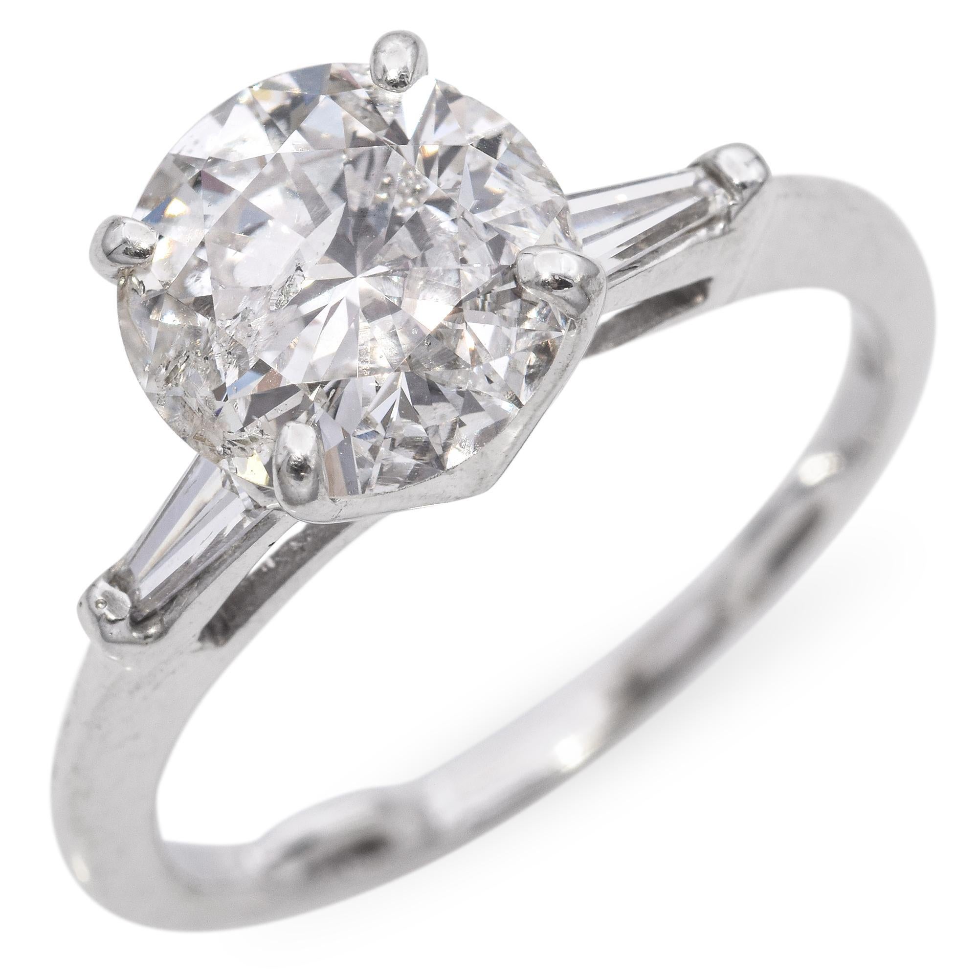 A stunning ring featuring a brilliant round-cut diamond at its center. The diamond is flanked by two tapered baguette-cut diamonds, adding elegance and symmetry to the design.
Set in a sleek white gold band with a four-prong setting that enhances