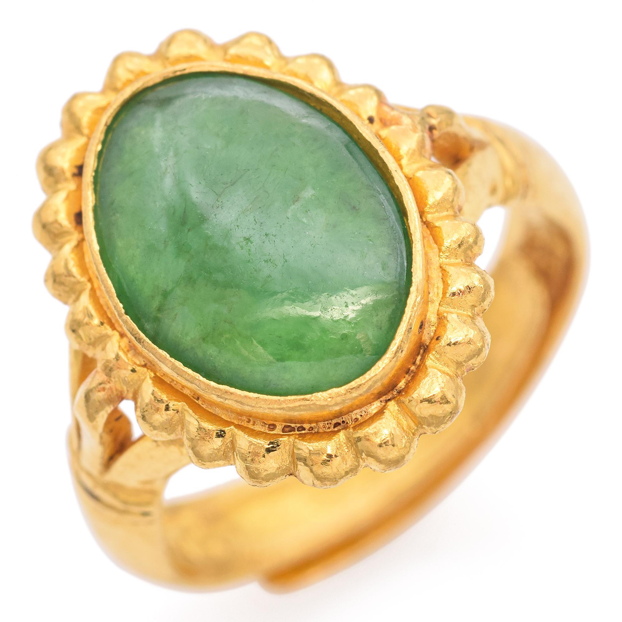 This striking antique ring is crafted in gold. It features a vibrant green oval Jade set in an ornate bezel. The gemstone is surrounded by a scalloped gold border, enhancing its rich color. The ring's band is intricately designed with detailed