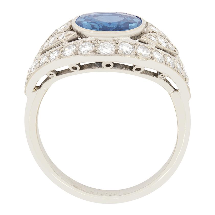Ein Cluster-Ring aus den 1950er Jahren, besetzt mit einem wunderschönen ovalen Saphir in einem kornmehlblauen Farbton, der von einem Cluster runder Diamanten im Brillantschliff umgeben ist. Der Saphir hat ein Gewicht von 2,70 Karat und wurde im