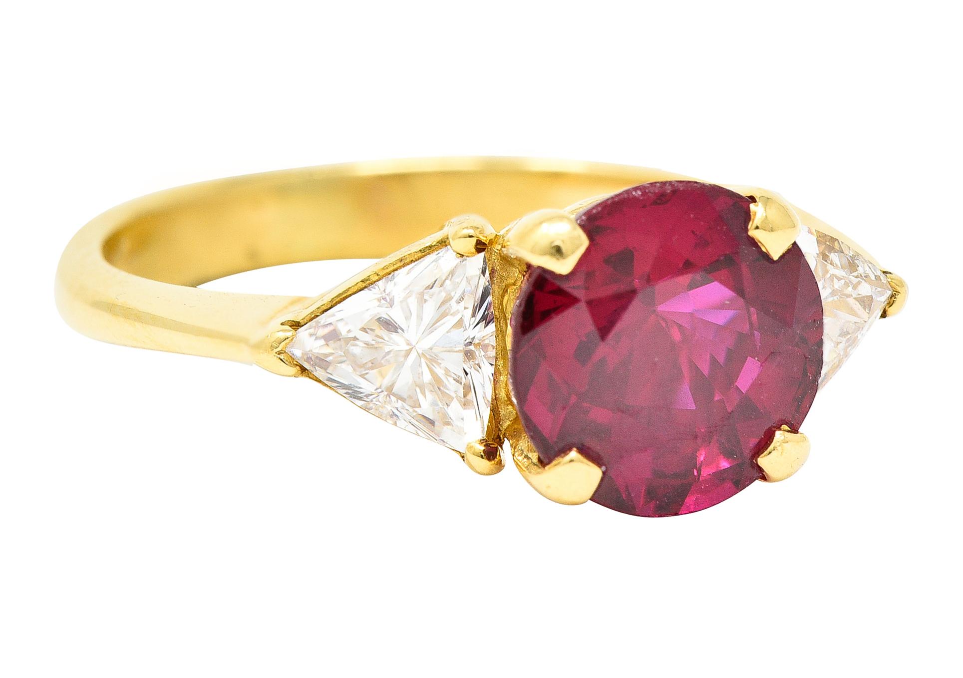 Three stone ring centers a basket set round cut ruby weighing approximately 2.10 carats. Transparent with vivid red color and no indications of heat - Thailand in origin. Flanked by trilliant cut diamonds weighing collectively approximately 0.68