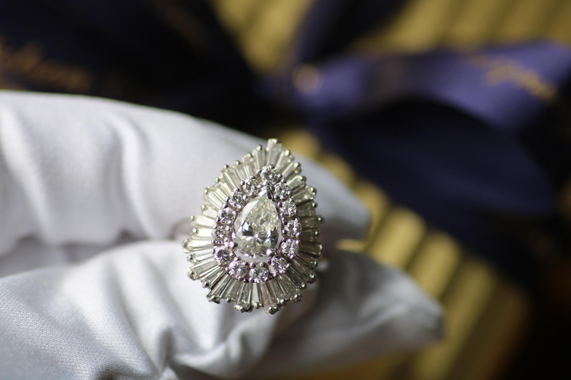 Un anello glamour con design da ballerina degli anni '70, realizzato in oro bianco 14 carati, con al centro un sorprendente diamante a forma di pera di circa 1,10 carati. La brillantezza del diamante centrale è accentuata da un'aureola di tredici