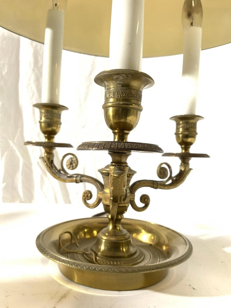 Antique French Napoleonic tole lamp has gold toned brass base and arms. Arms have ornate s curve structures. Each arm has candlestick style bulb socket at ends. Top of lamp has eagle finial, arrow & tail structure underneath. 
Search terms: Lamp,
