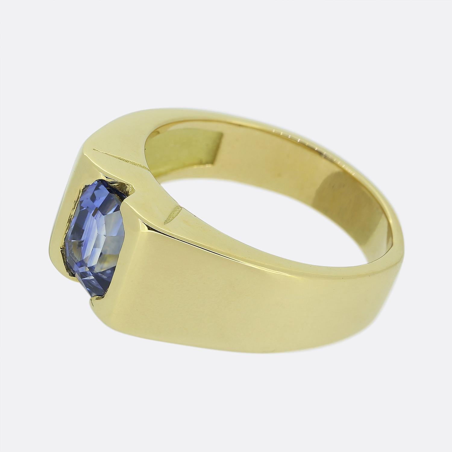 Here we have a sleek 18ct yellow gold solitaire ring. At the centre of the face we find a single emerald cut sapphire possessing a striking natural blue colour tone, securely semi bezel set amidst a pair of wide square shoulders. The design is