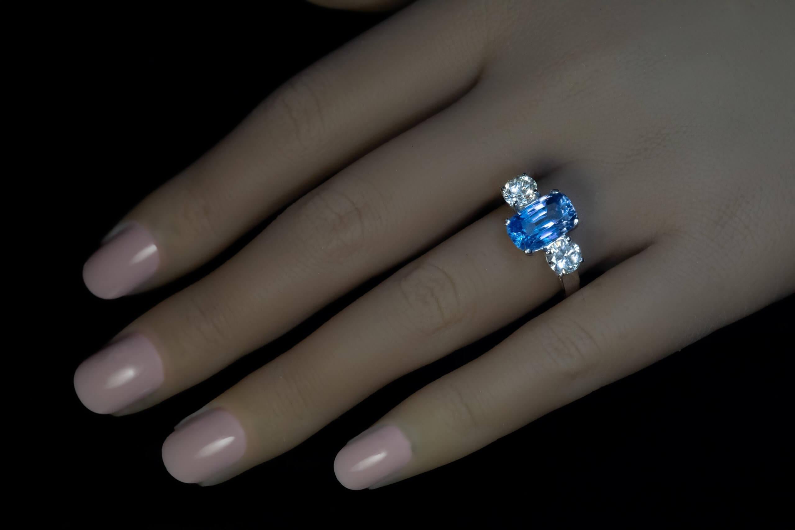 Este anillo de compromiso de tres piedras está elaborado en oro blanco de 14 quilates, con un zafiro de Ceilán sin calentar, talla cojín, de 3,92 quilates. El zafiro muestra un vivo color azul violáceo, excelente brillo y es limpio a la vista.  La