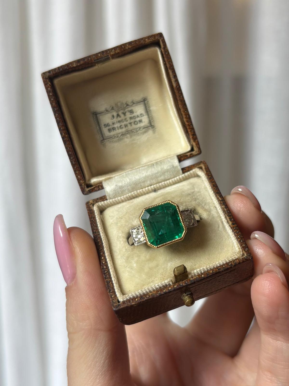 A truly stunning vintage emerald and old cut diamond three stone ring set in platinum and 18k yellow gold. 

The ring is predominately set with a large 4.16ct vibrant emerald cut emerald, securely encased in a mille grain yellow gold setting. The
