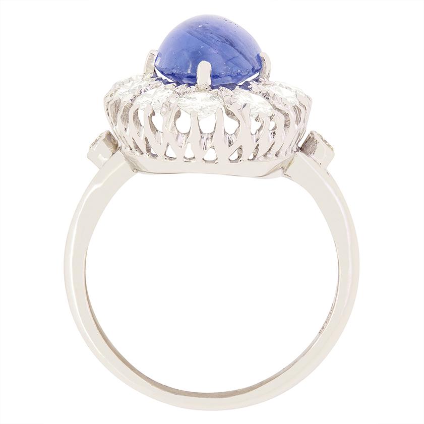 This beautiful cluster ring dates to the 1950s and showcases a stunning 5.00 carat cabochon cut sapphire at its centre.. The light blue sapphire has an ethereal quality in its completely smooth oval shape. Encircling the sapphire are a halo of round