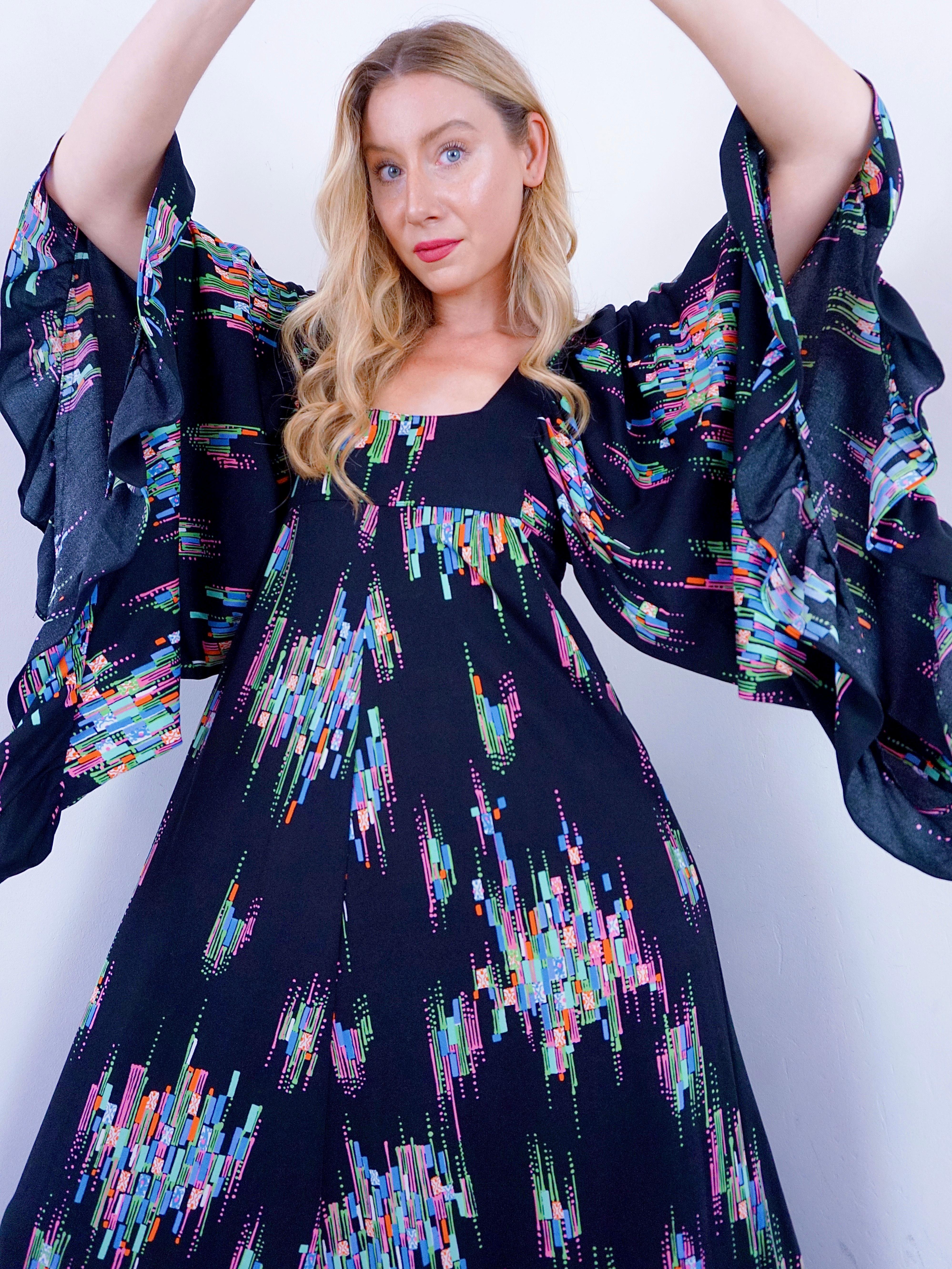 Incredible 1970s angel sleeves maxi dress. She has a black base with a rainbow coloured geometric print all over. A square neckline with a waterfall maxi coming from under the bust finished with a ruffled hemline that matches her ruffled