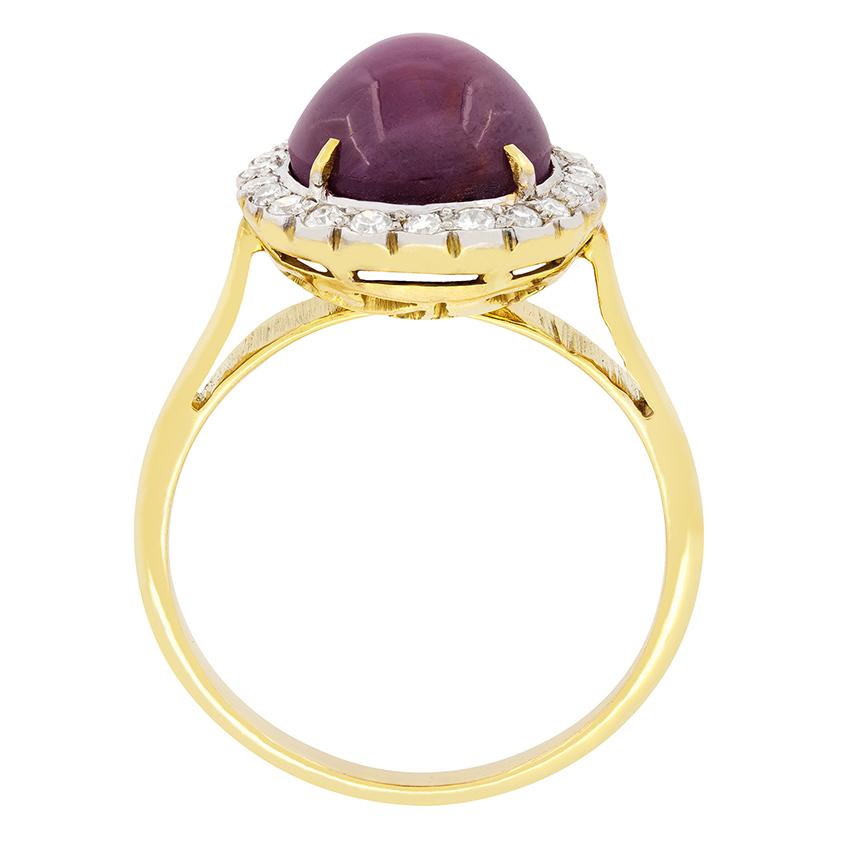 This awe inspiring vintage ring features at its center a mesmerizing star ruby. The gem stone is a 7.75 carat cabochon cut stone with an unusual purple-red colour. It is claw set within 18 carat yellow gold and surrounded by a halo of eight cut
