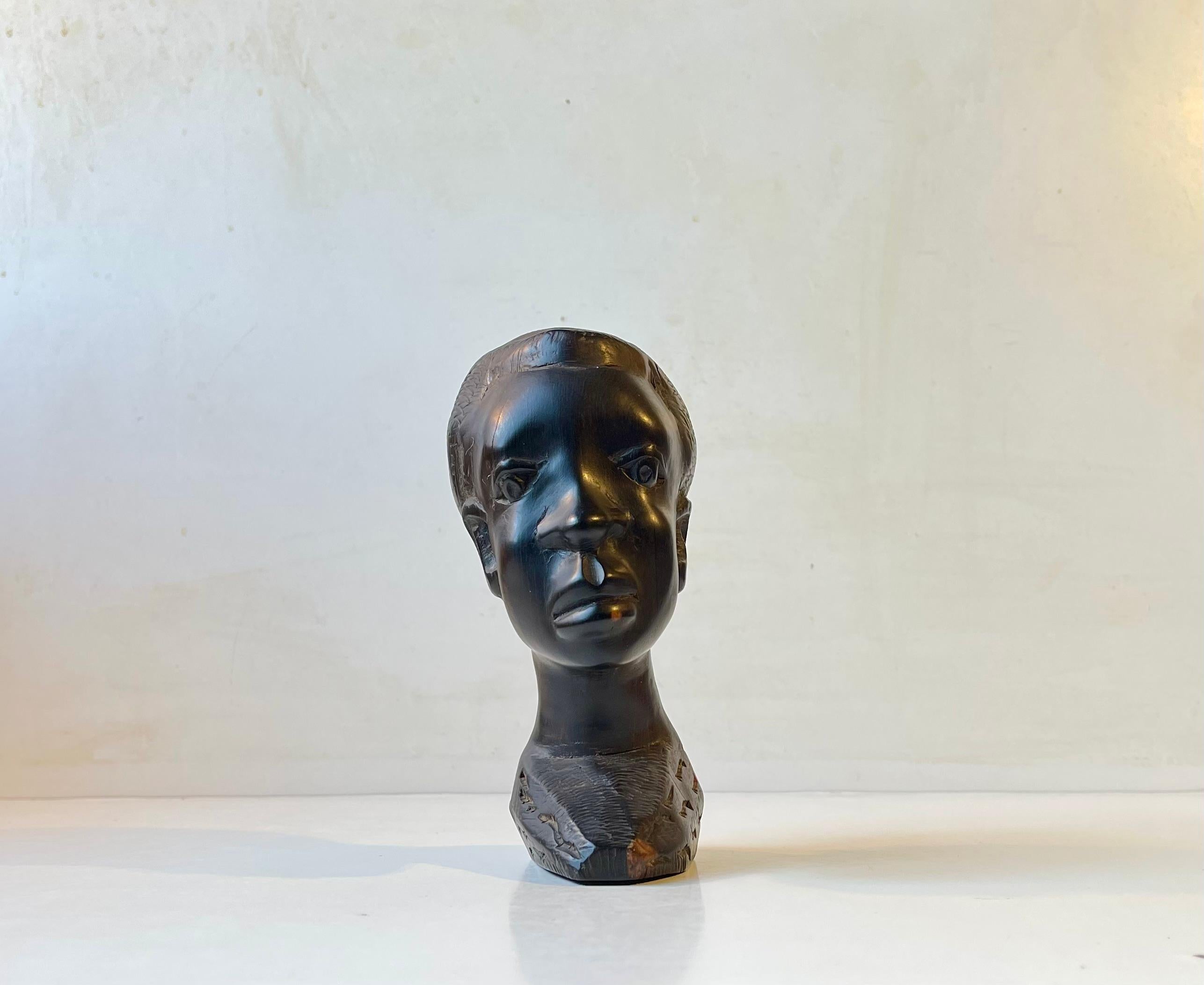 Small scaled african head bust hand carved in black wood. It has a slight tilt - leaning to one side. It features fine details and textures. Particularly its cheekbones and hair. It was made in Africa at the latest during the 1960s where it was