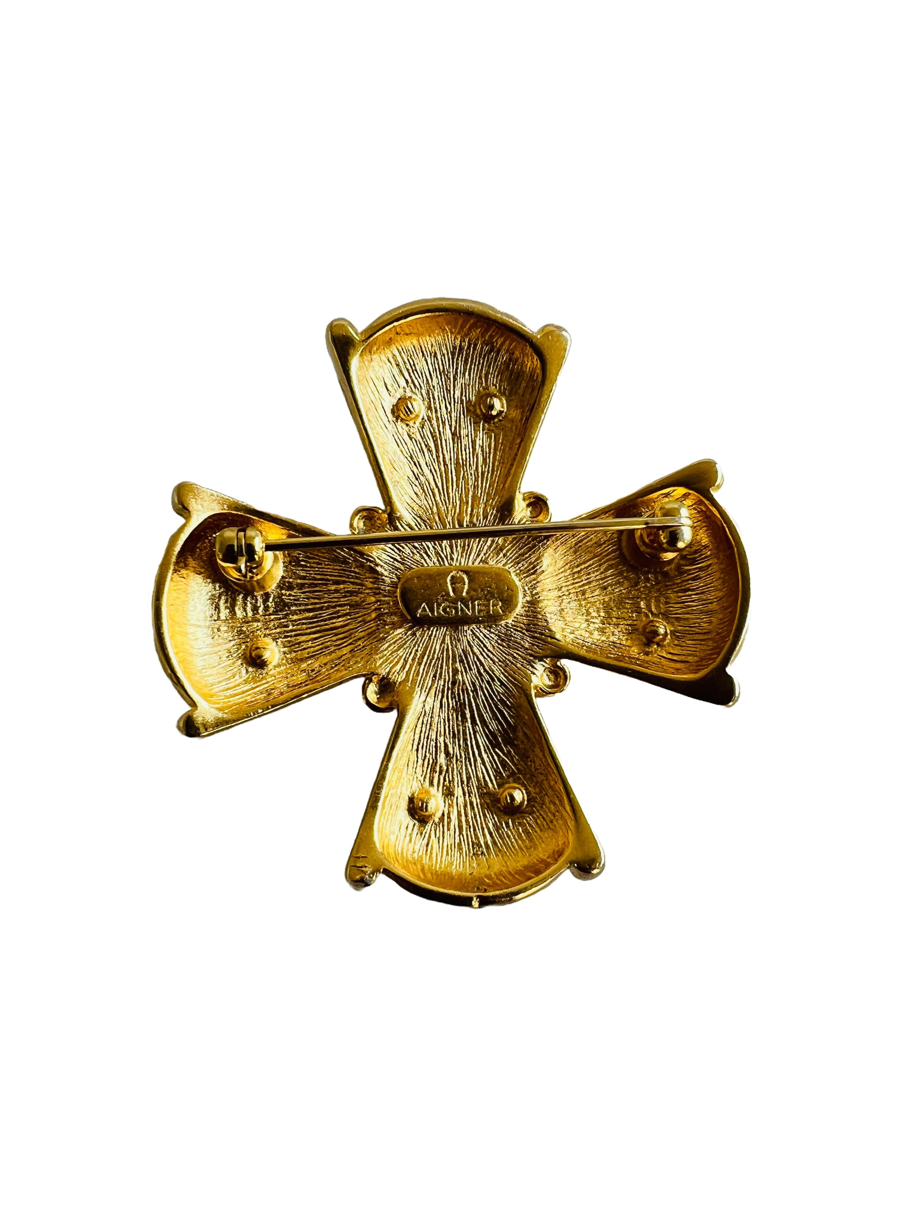 This Aigner brooch is a stunning tribute to timeless elegance, featuring a bold Maltese cross design in rich gold-tone metal with a striking red sugarloaf cabochon centerpiece. Each arm of the cross is marked with Aigner's signature 