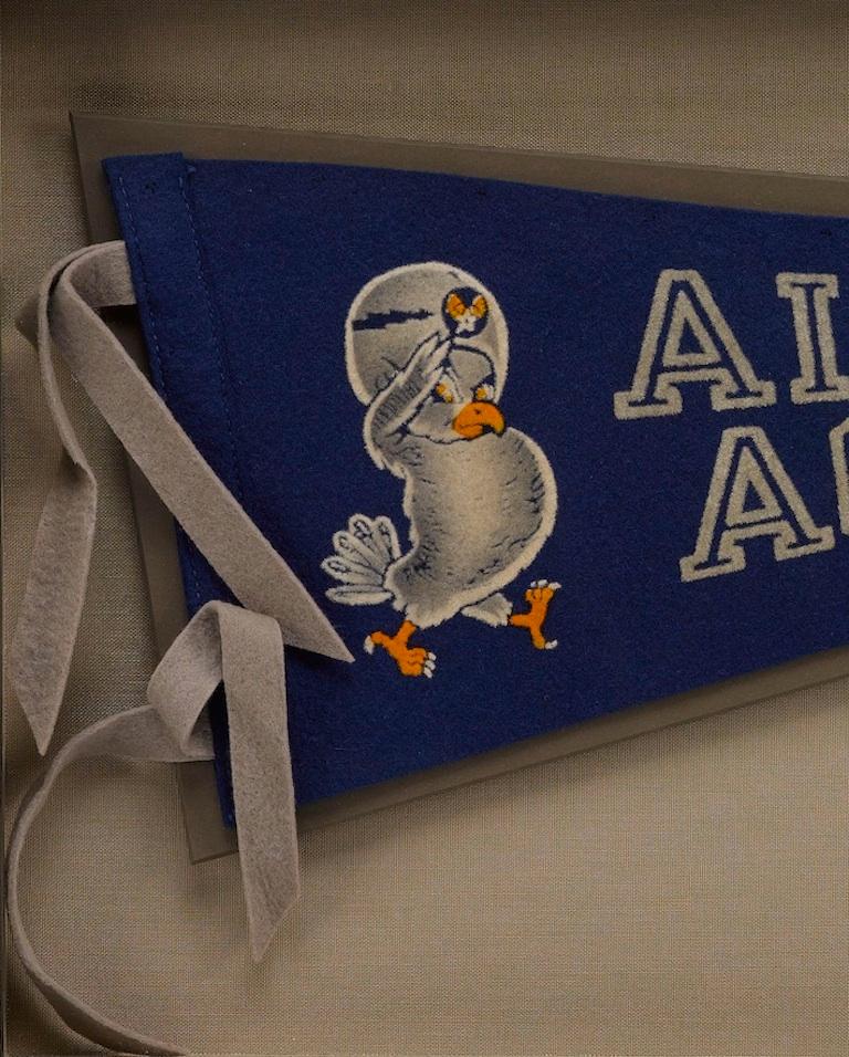 This vintage Air Force Academy pennant dates to the 1960s. The felt pennant has a blue background with a matching headband and gray ties. In white outlined letters, the pennant reads, “Air Force Academy.” Just to the left is a saluting falcon
