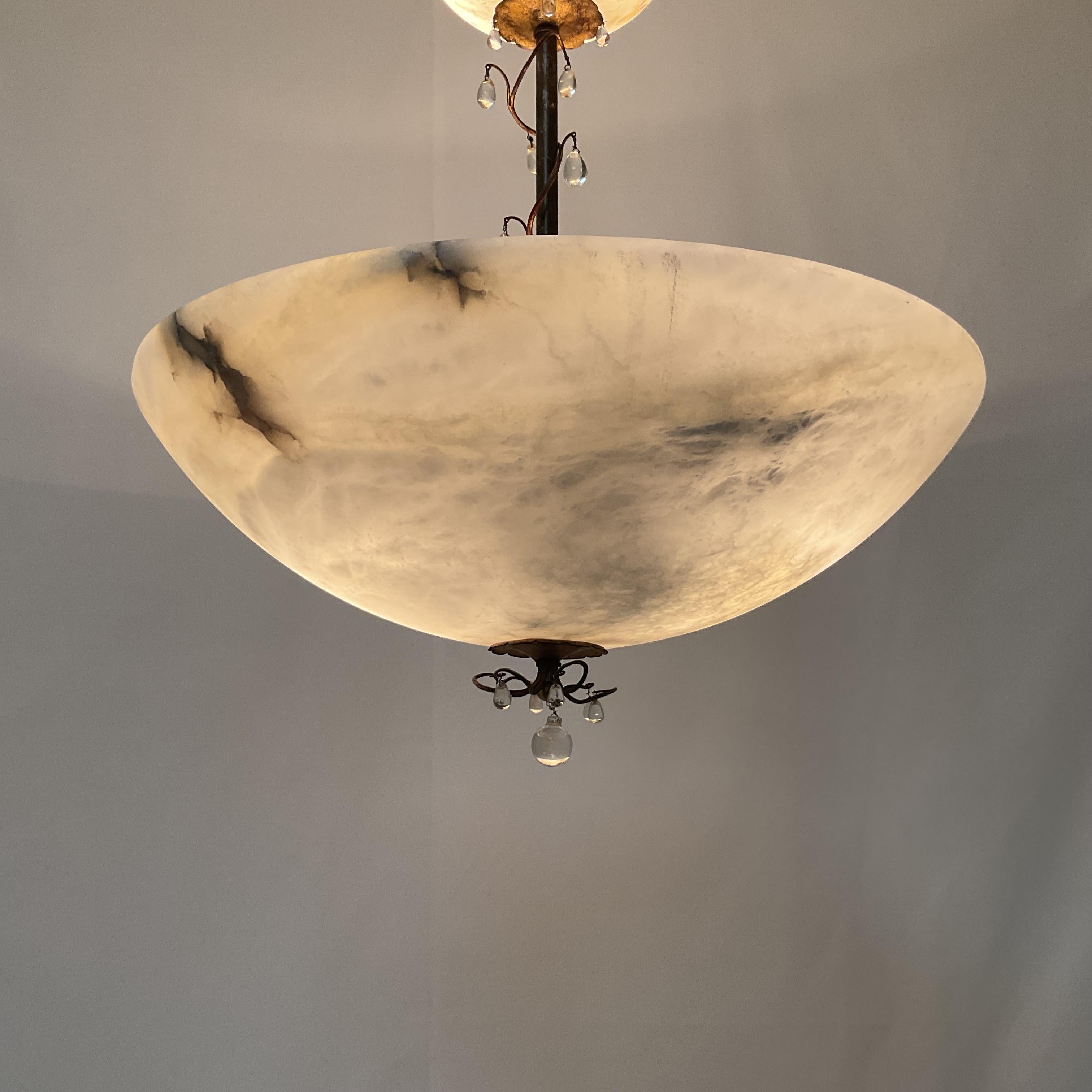 Questo elegante lampadario vintage è caratterizzato da una ciotola di alabastro intagliata a mano sospesa su uno stelo decorativo in metallo ornato da delicati accenti di cristallo a goccia. Le venature naturali dell'alabastro creano una luce calda