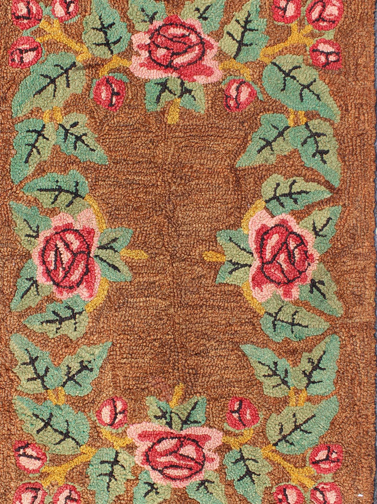 This colorful American Hooked rug depicts a variety of vines and blossoming flowers in an assortment of colors extending throughout the light orange and tan background. The colors in the flowers are marigold, rose, pink, greens and