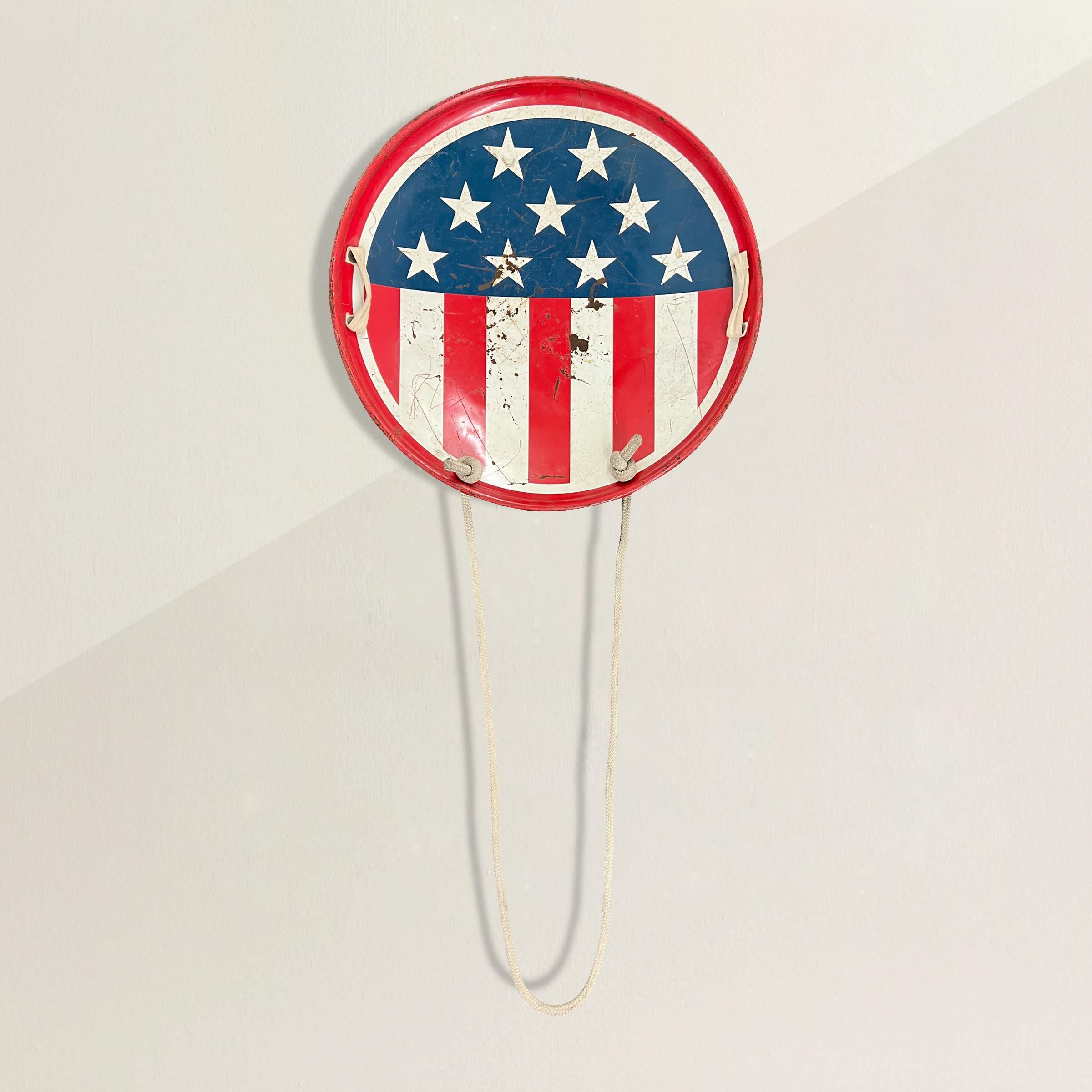 Une charmante et ludique soucoupe-luge américaine vintage peinte avec un motif de drapeau américain, et conservant ses poignées et sa corde de traction d'origine. Nous avons fait fabriquer un support en acier sur mesure pour que vous puissiez