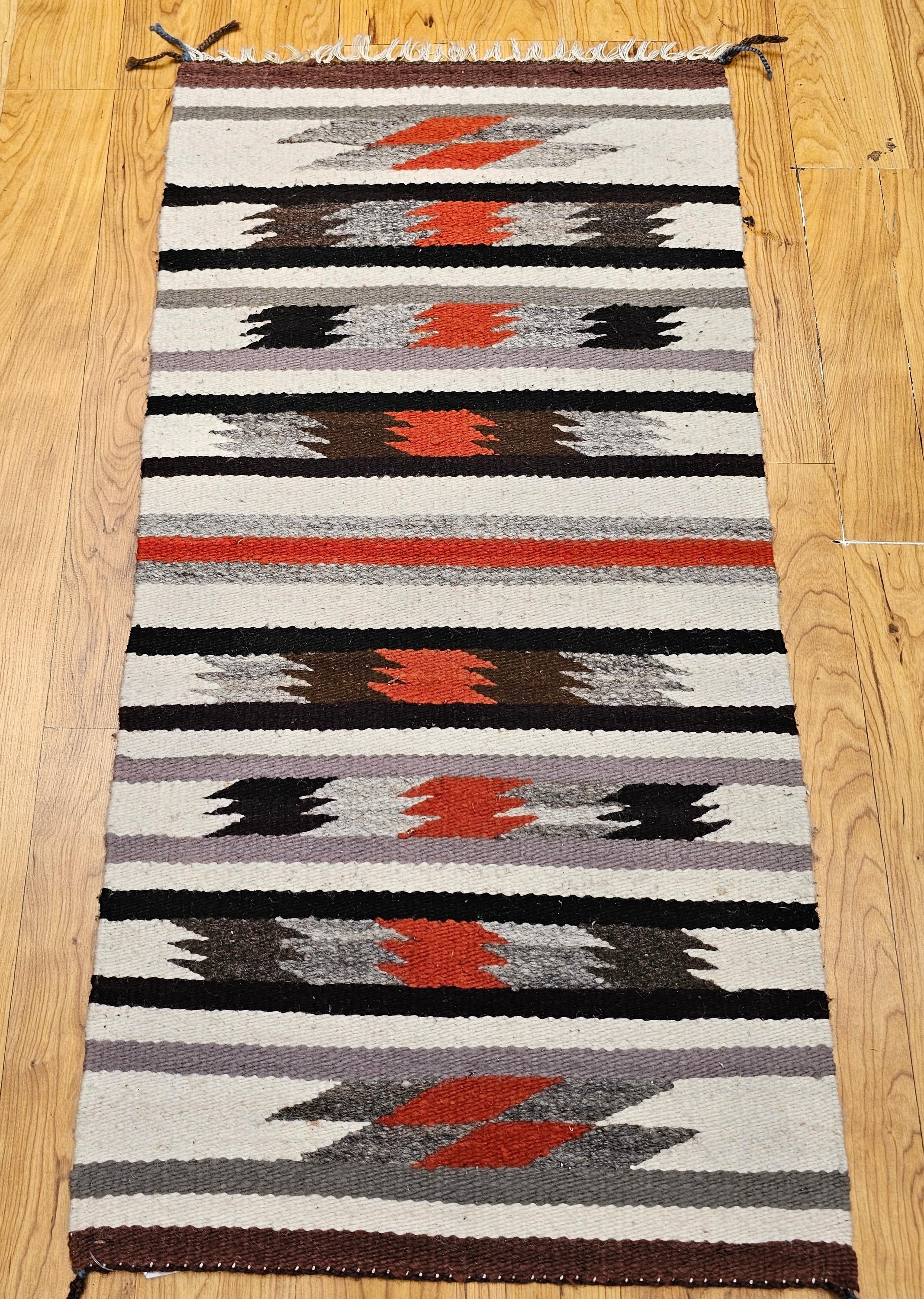 This vintage American Navajo rug features a striking striped design in a harmonious blend of earthy Southwestern tones, including ivory, black, gray, and rust-red. Woven in the mid-20th century by a skilled Diné (Navajo) artisan, this piece reflects
