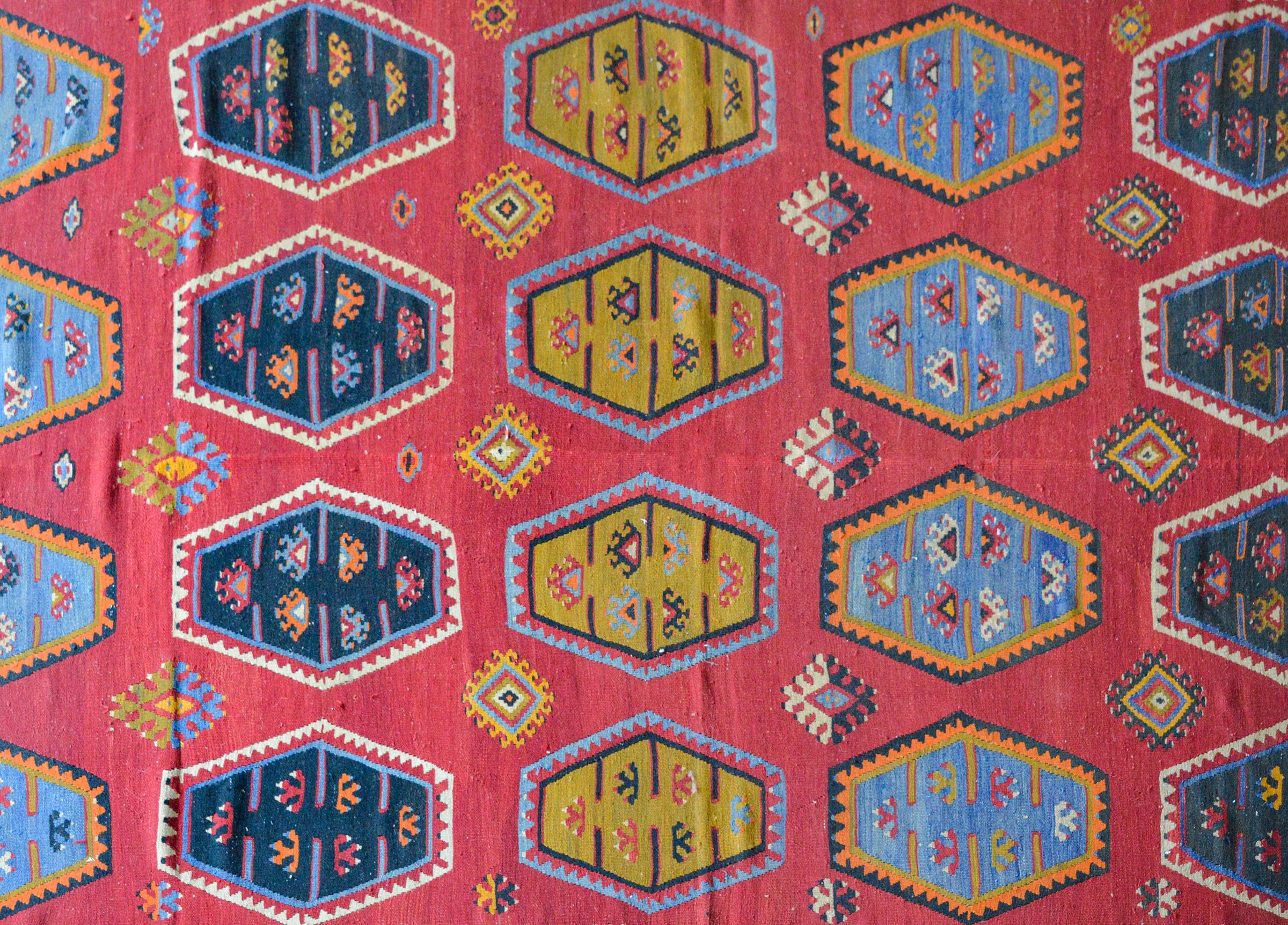 A wonderful mid-20th century Anatolian Turkish kilim rug with several hexagonal medallions each with stylized flowers and woven in light and dark indigo, green, orange, and white against a solid crimson background. The border is bold with two