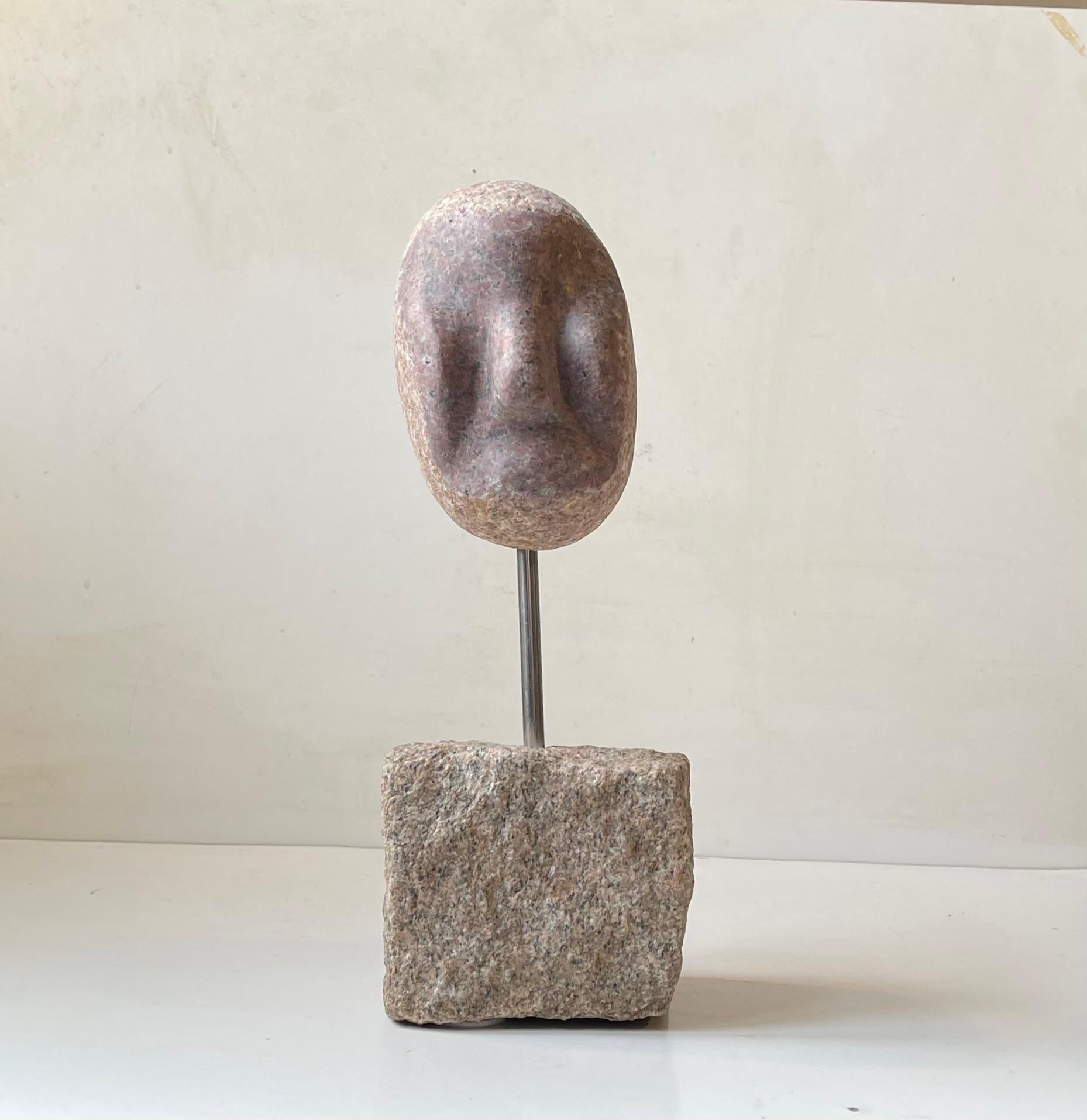 Cabeza de piedra andrógina sin expresión y base ejecutadas en granito tallado. Duro y suave en expresión y forma al mismo tiempo. Mientras que la cabeza está realizada en un granito rosa/rojo, la base parece ser un adoquín modificado que sujeta la