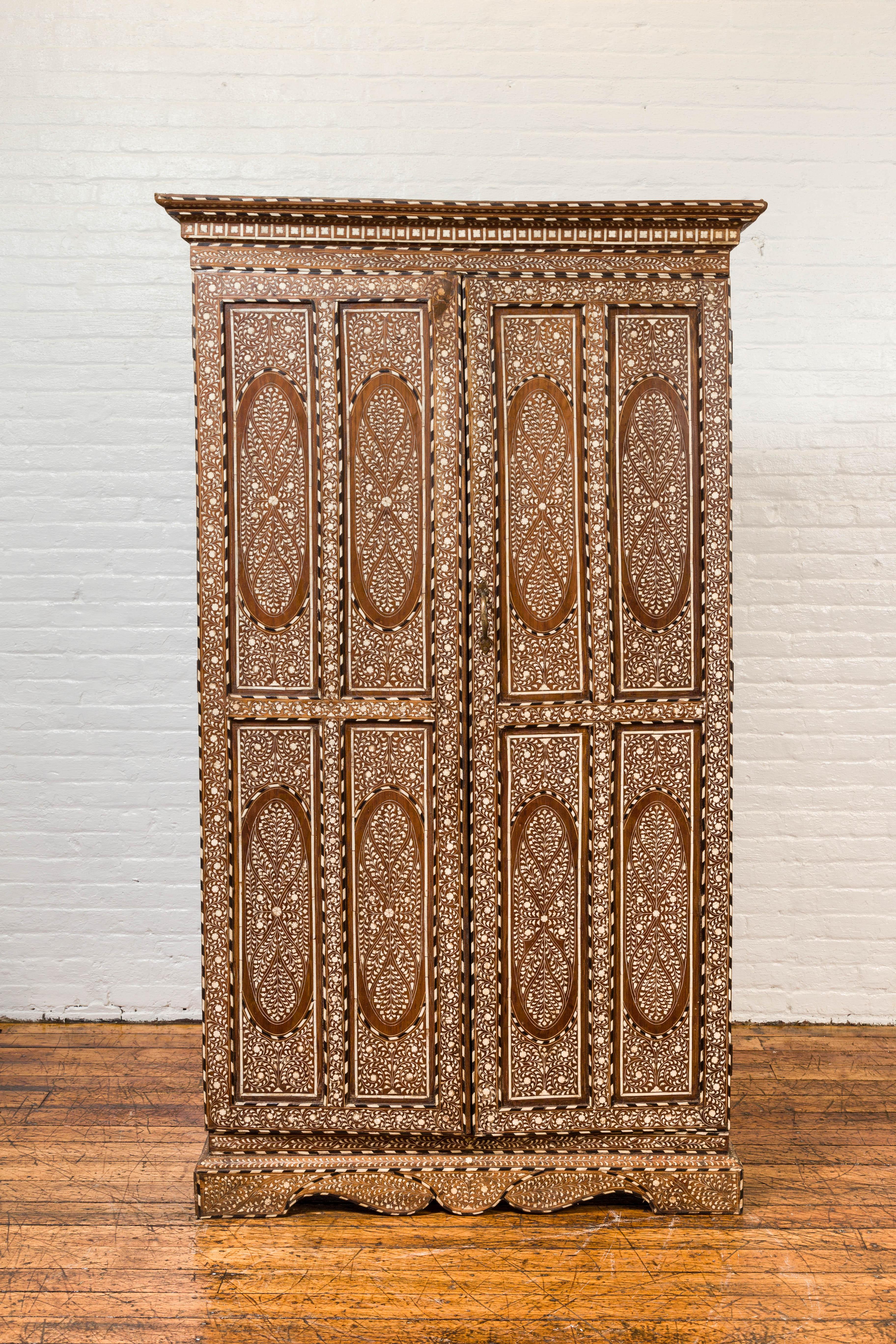 A vintage Anglo-Indian inlaid wooden wardrobe cabinet from the mid-20th century, with ebonized accents, bone and horn inlay. Capturing our attention with its intricate decor, his tall cabinet is adorned with an abundant décor of foliage and