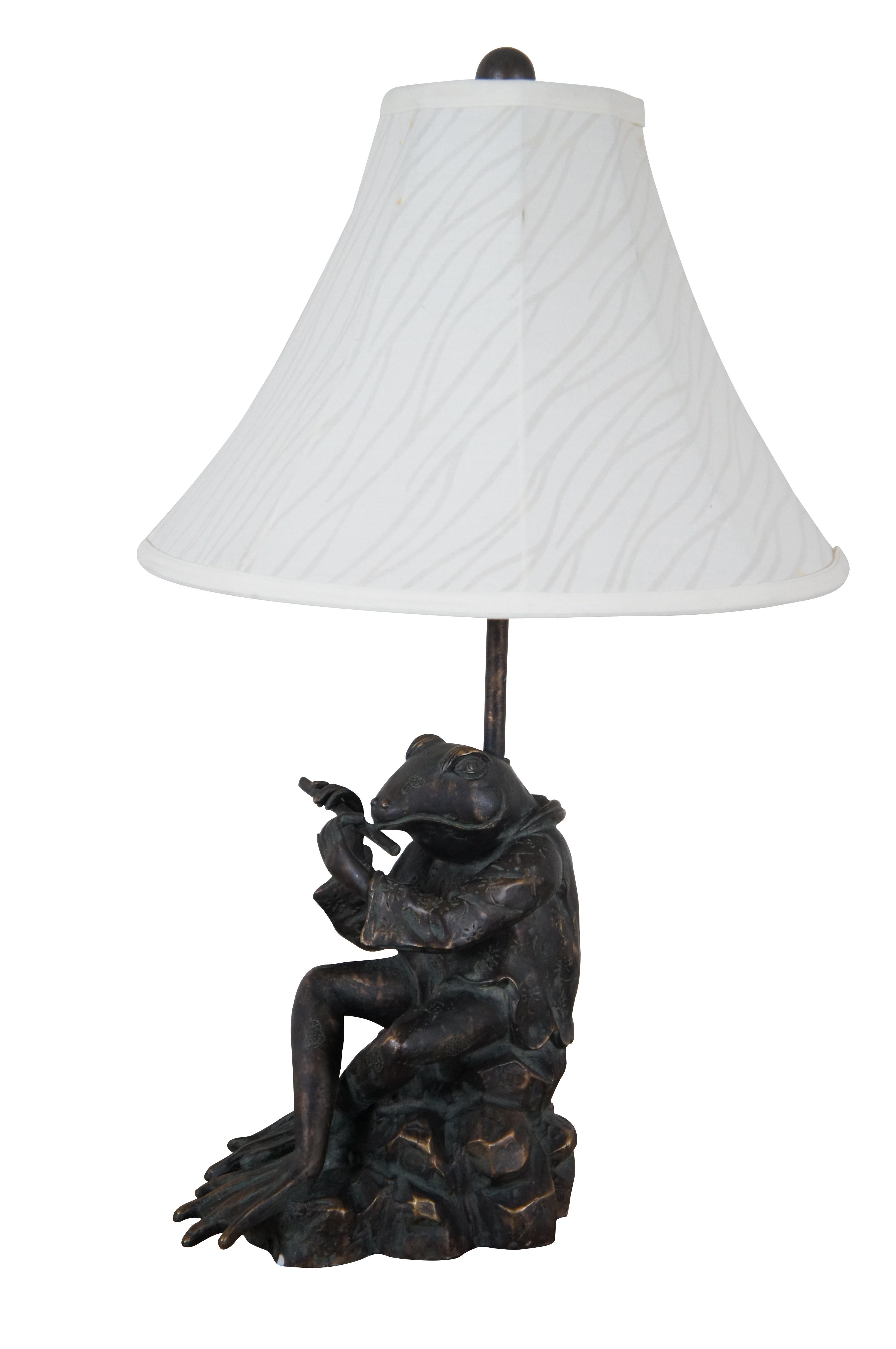 Sculptural composition table lamp in  the shape of a bronzed anthropomorphic frog, dressed in a short patterned Japanese kimono jacket and playing a flute.  Includes a white zebra patterned cloth shade and wood ball finial.

Dimensions:
9.75