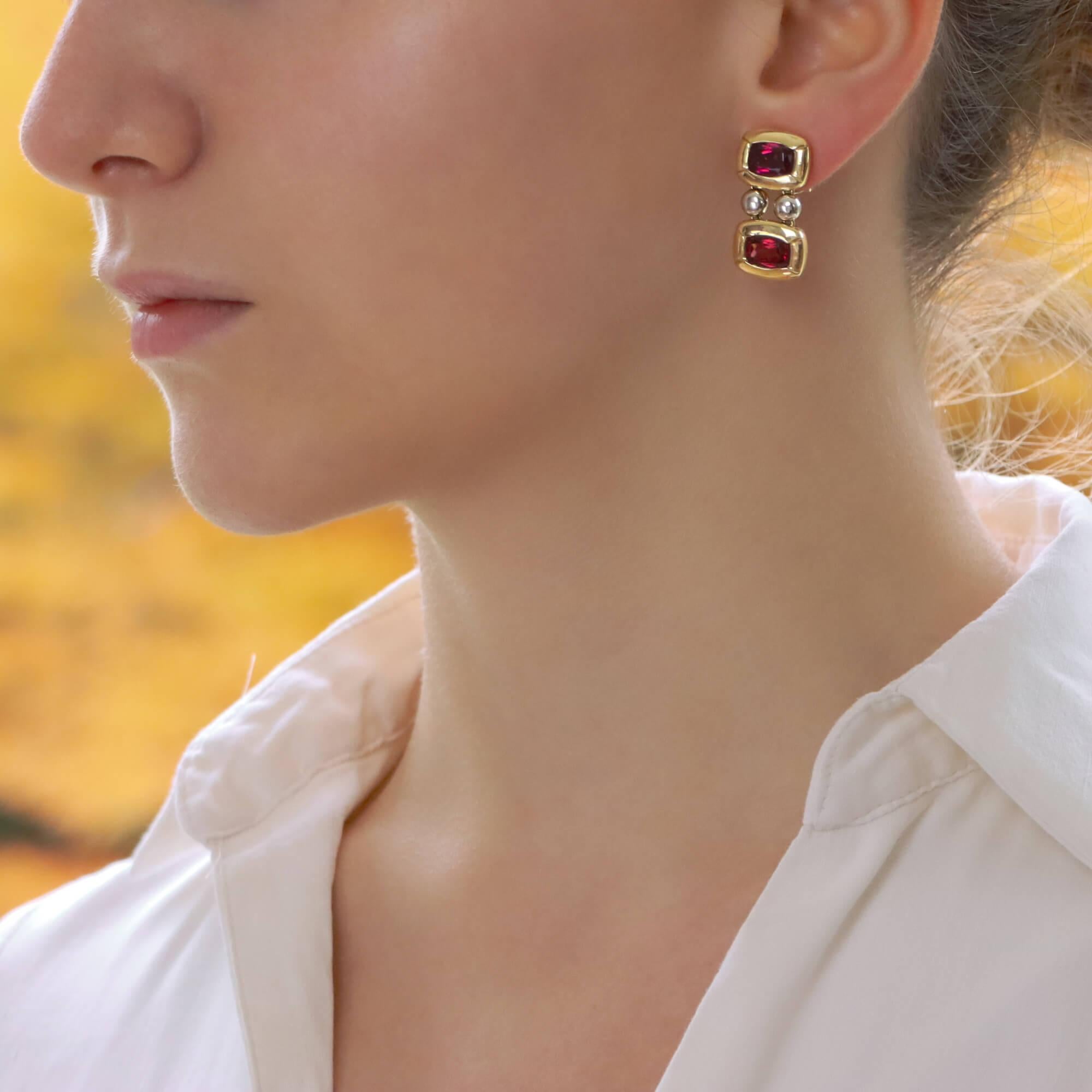 A beautiful pair of signed Antonini garnet drop earrings set in 18k yellow and white gold.

Each earring is composed of two vibrant red garnets, which are rubover set in a rectangular panel setting. The panels are connected by two white gold spheres